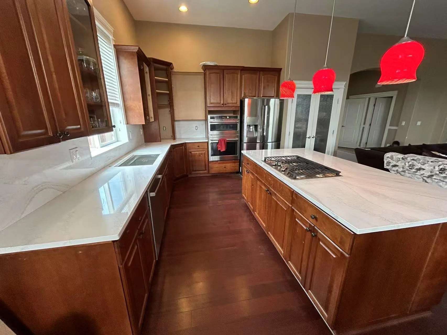 A spacious kitchen with wooden cabinets, a large island with a white countertop and red pendant lights above, stainless steel appliances including a refrigerator, oven, and microwave, and a dark hardwood floor.