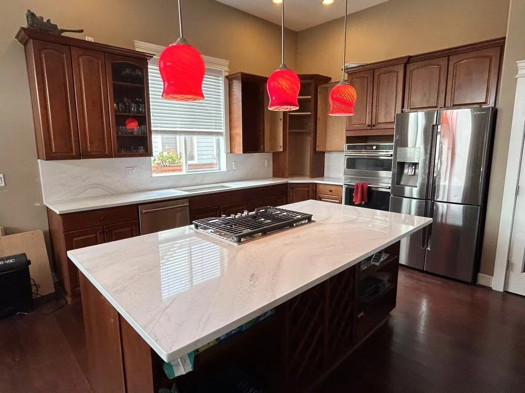 Modern kitchen with dark wooden cabinets, a white marble countertop island with a built-in stove, three red pendant lights hanging above the island, a stainless steel refrigerator, and a window with white blinds over the sink.