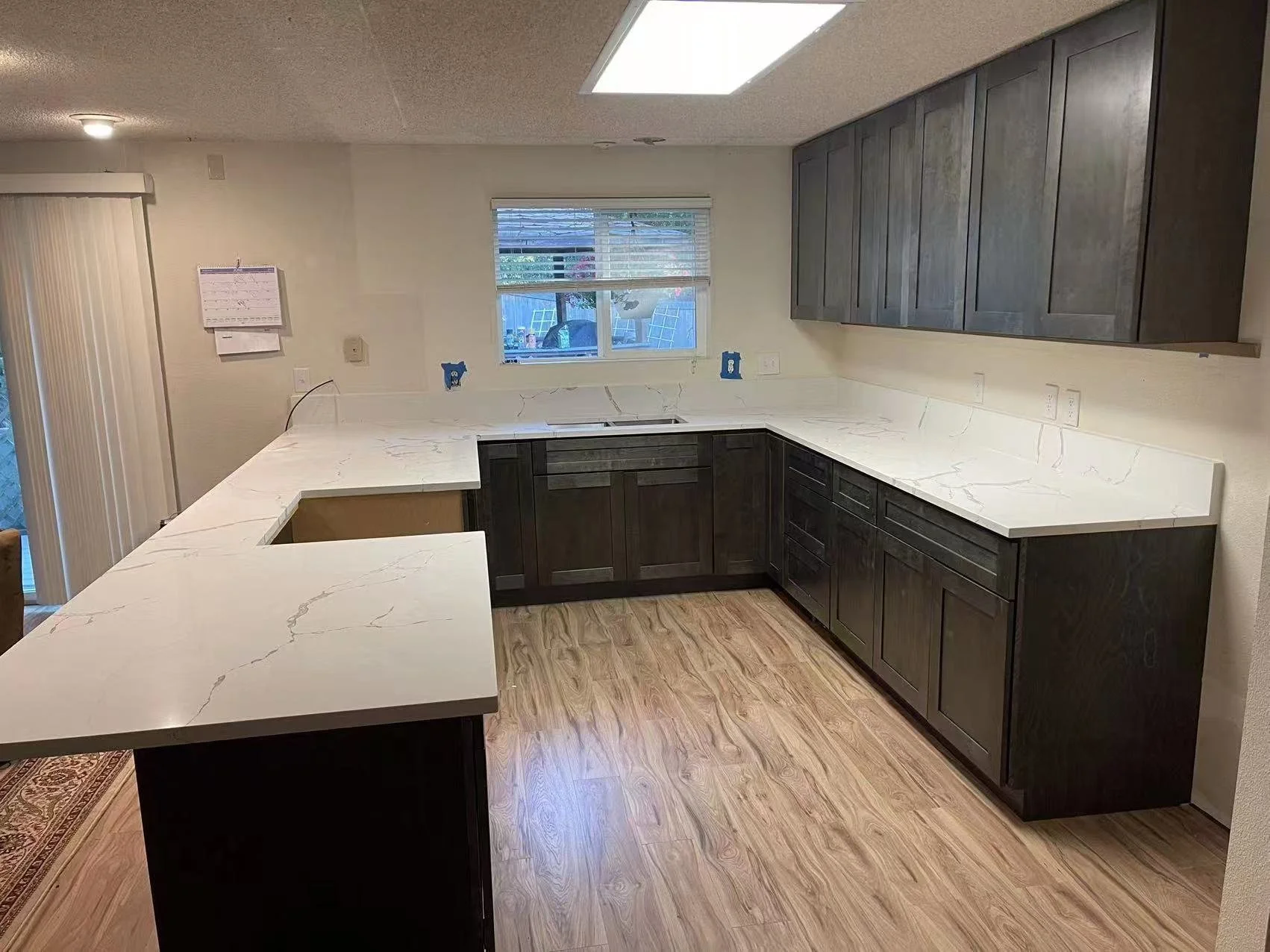 A kitchen with dark wooden cabinets and white marble countertops, a window with a view outside, and a sliding glass door to the left.