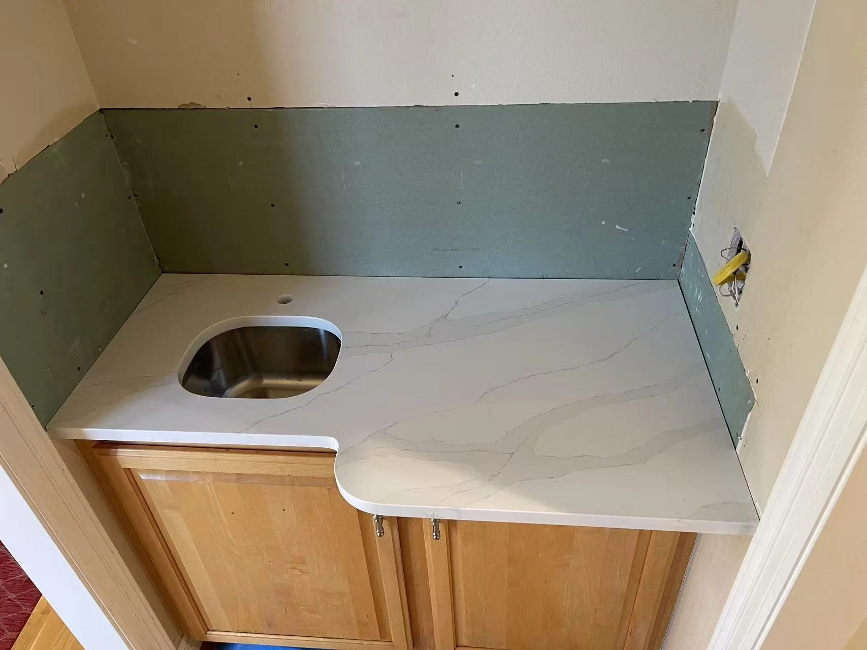 A small kitchen countertop with a built-in stainless steel sink, wooden cabinet beneath, and a green drywall backsplash; a hole in the wall for an outlet is visible on the right.