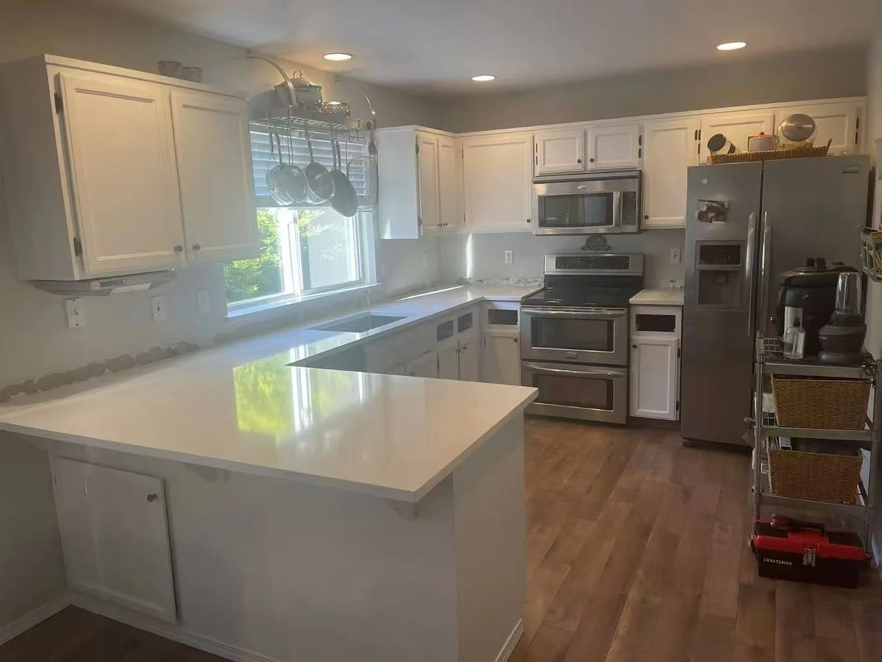 Modern kitchen with white cabinets, stainless steel appliances, wooden flooring, window with blinds, hanging pots and pans, and a small rolling storage cart.