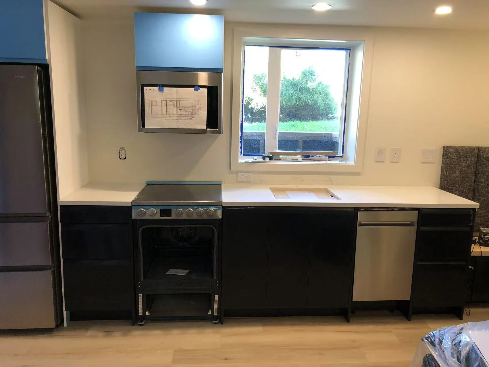 Empty kitchen with a refrigerator on the left, a stove in the center, and a dishwasher on the right, with a window above the countertop showing a grassy yard outside.