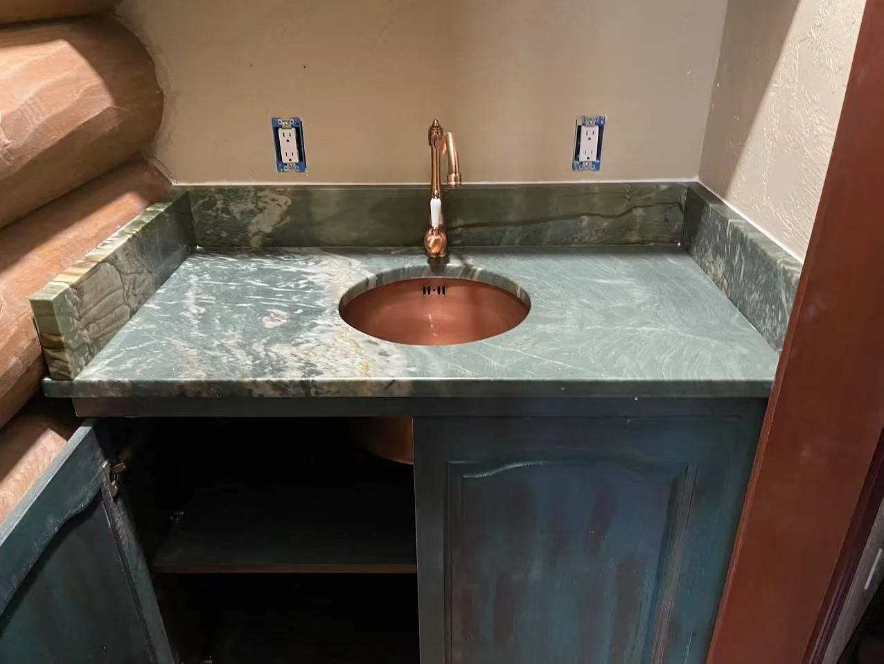 A green marble countertop with a copper sink and matching copper faucet, mounted beneath two electrical outlets, with unfinished wall and cabinet space below.