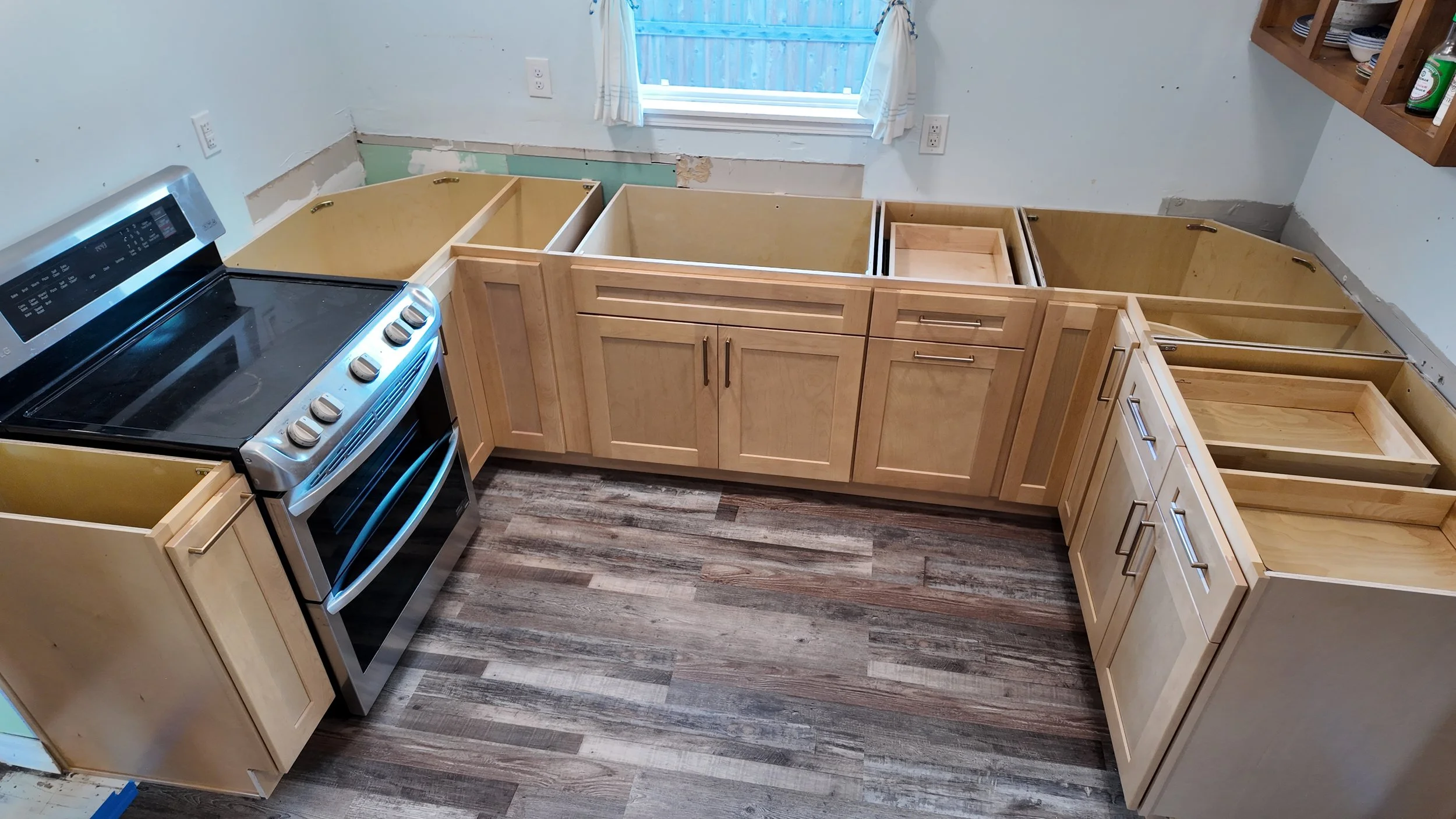 Kitchen under renovation with unfinished wooden cabinets, a black oven, and a window with curtains.