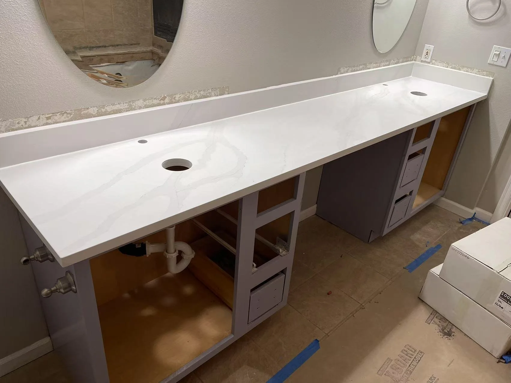 Under construction bathroom vanity with a white marble countertop, two pre-cut sink holes, and unfinished cabinetry, with plumbing pipes visible underneath.