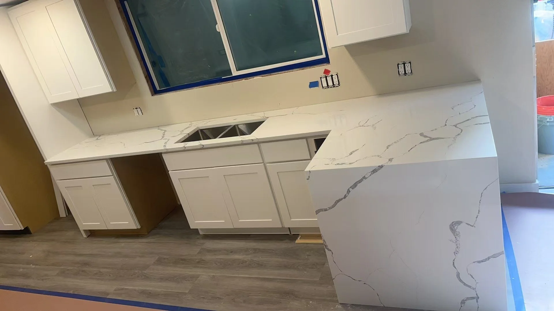 Kitchen with white cabinetry, marble countertops, and a window above the sink. The cabinetry and countertop are not yet finished, and the floor is dark wood.
