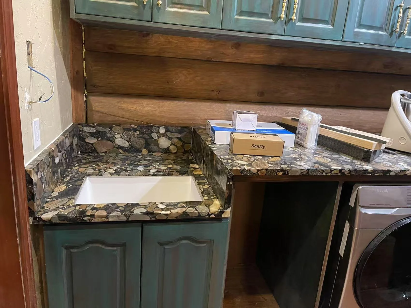 A partially installed kitchen countertop with a pebble stone pattern, a square white sink, and various construction materials on the counter. There are green cabinets, wood paneling on the wall, and a washing machine visible.