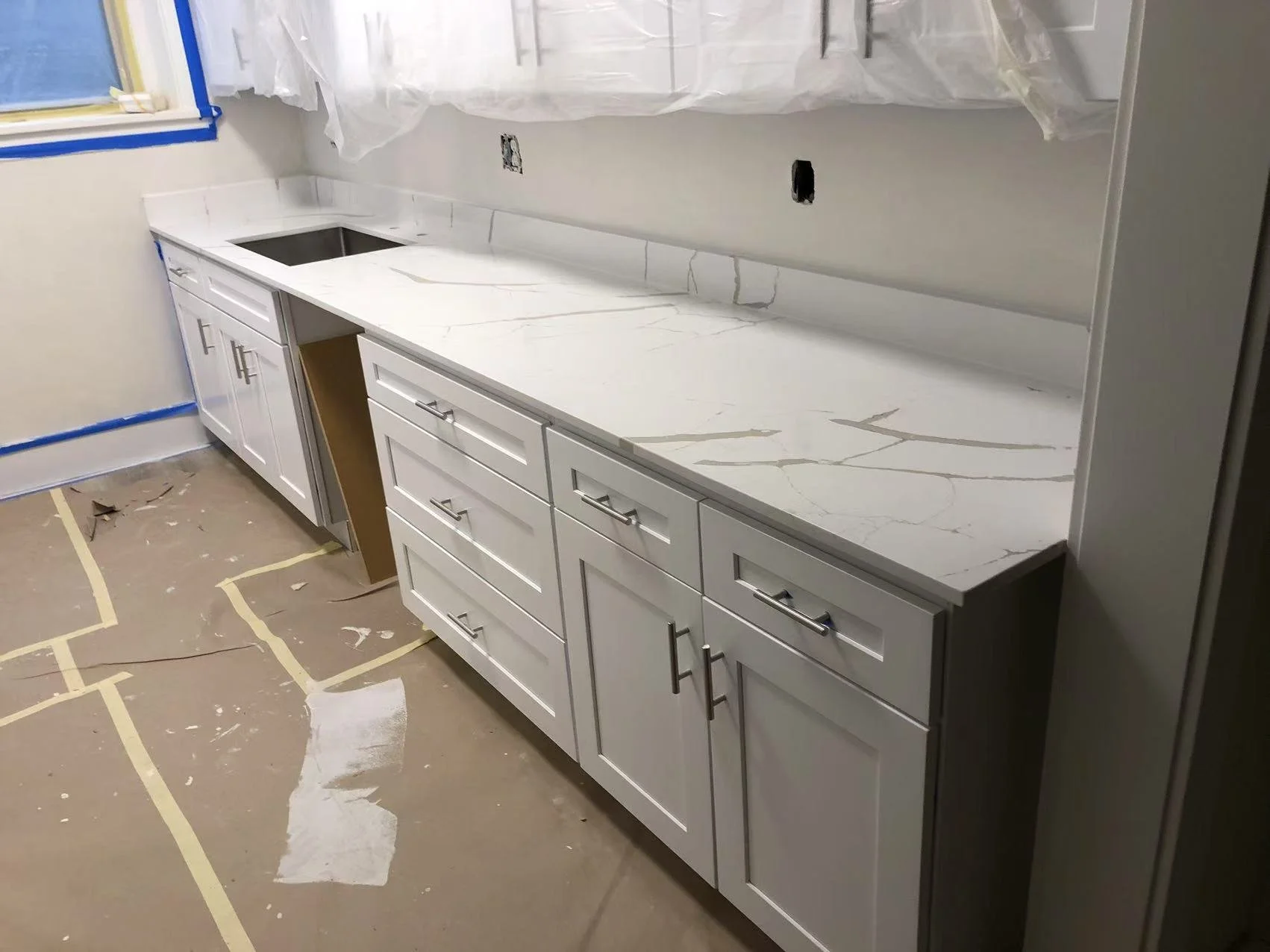 Kitchen countertop installation in progress with white cabinets, marble-like countertop with gold veining, and a cutout for a sink. Protective plastic and painter's tape are visible, indicating ongoing renovation.