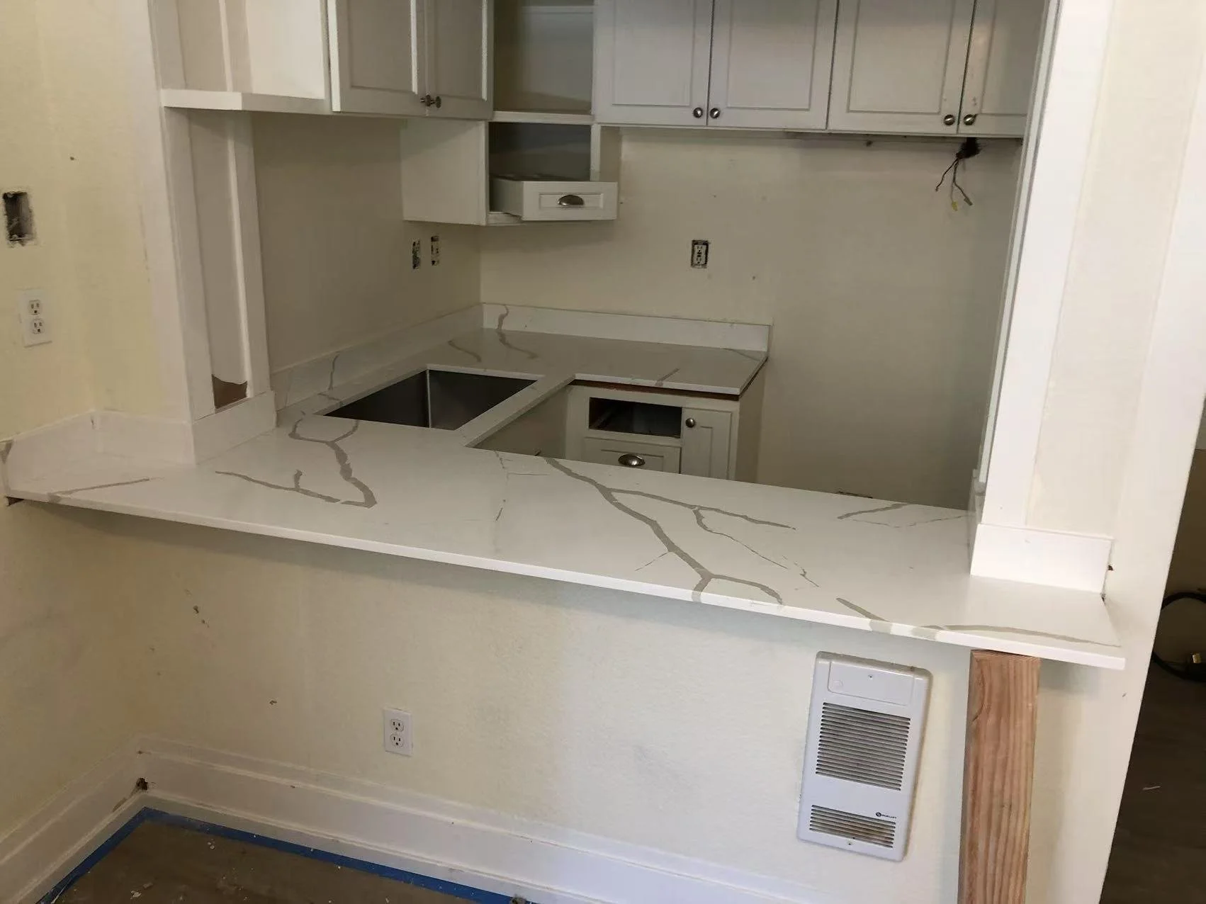 Kitchen with white cabinets, marble countertop, and a small open section of upper cabinets. The sink is missing, and electrical outlets are visible on the wall. There is a cutout in the wall for a stove or oven, and some wiring is exposed. A vent is 