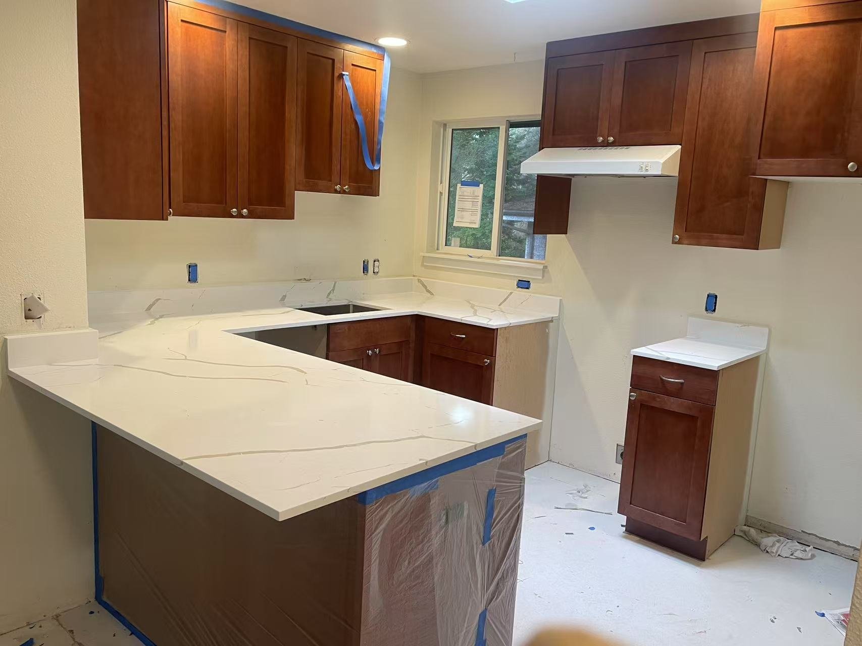 Kitchen under construction with wooden cabinets, white marble countertops, and a window above the sink area.