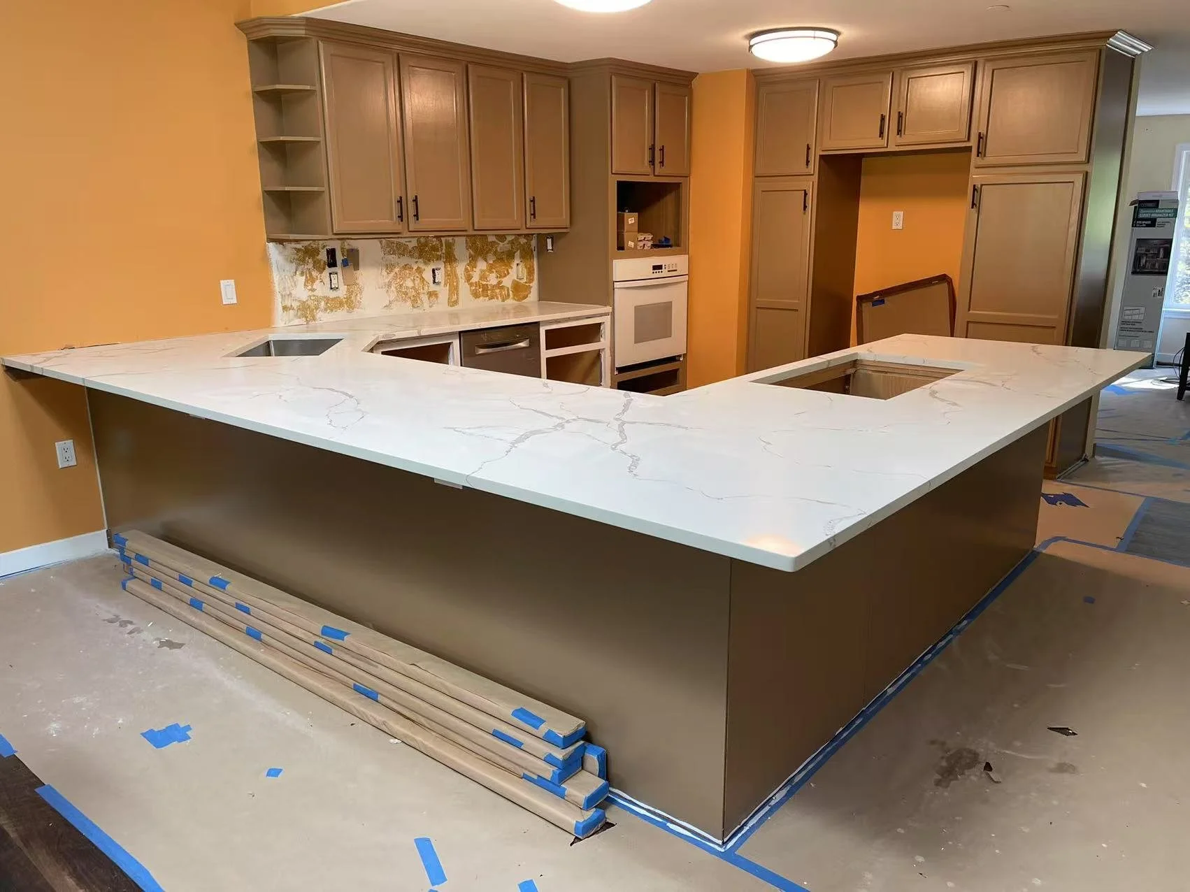 Kitchen under renovation with a large marble countertop island, wooden cabinets, and appliances, with construction materials and blue painter's tape on the floor.