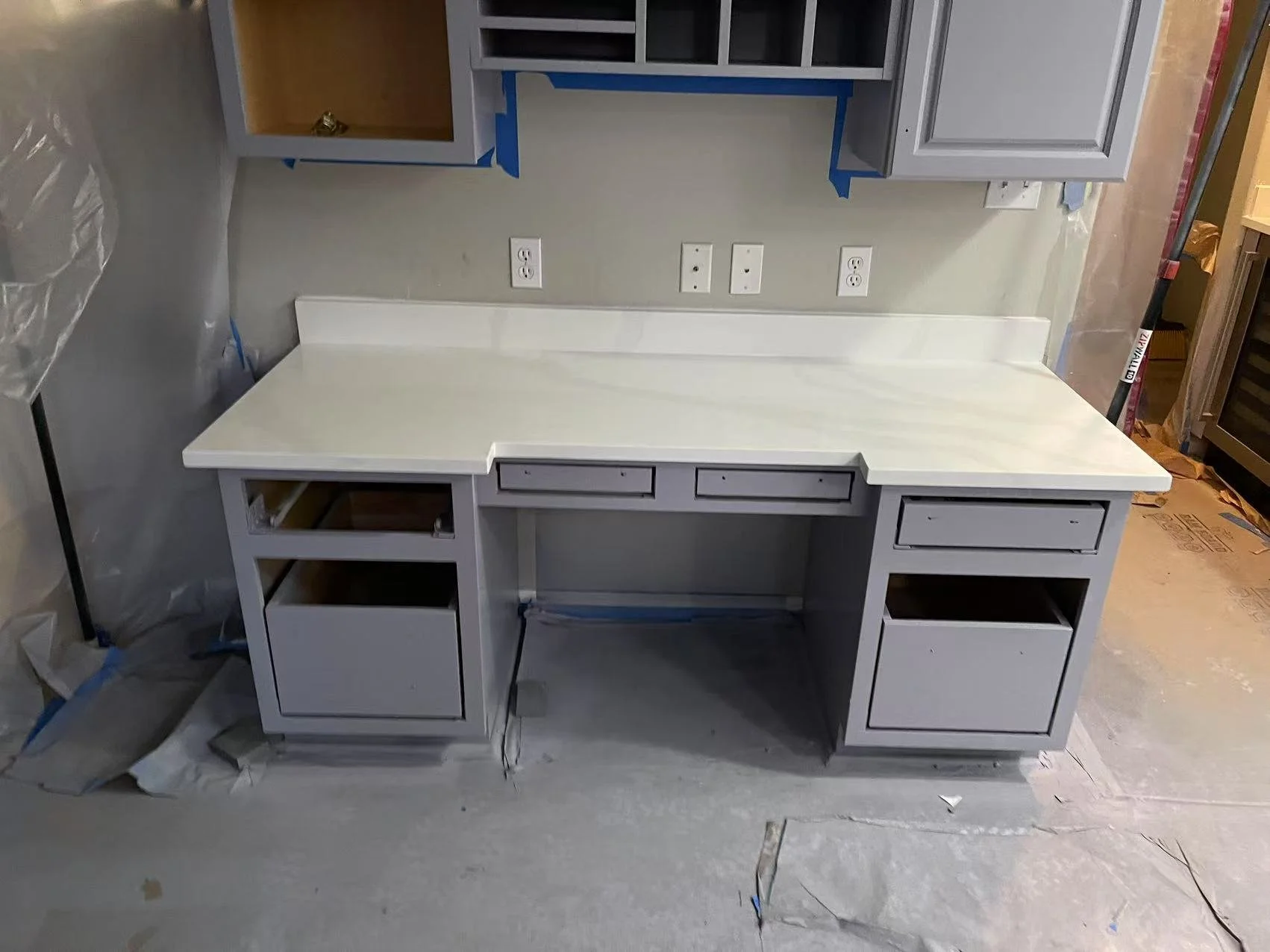 Gray built-in desk with open drawers and cabinets under construction, with construction materials and tools nearby in a room with electrical outlets and wall painting in progress.
