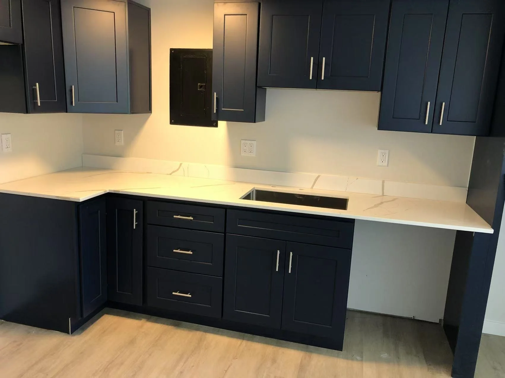 Kitchen with dark blue cabinets, white marble countertop, and a built-in black sink. The wall has multiple electrical outlets, and there is a black access panel on the wall.