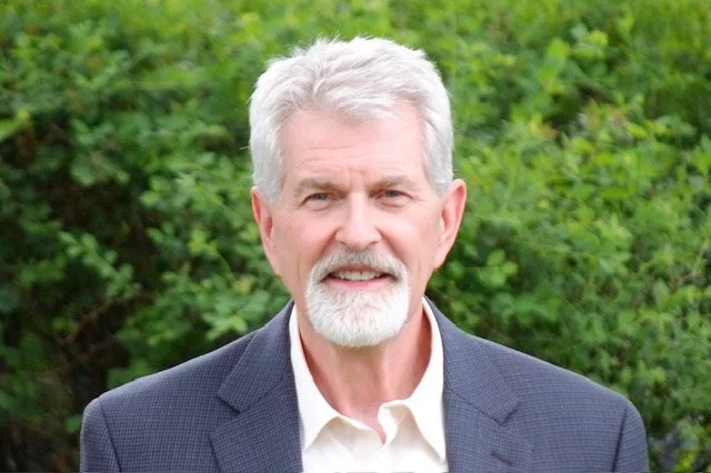 Portrait of an older man with gray hair, a beard, dressed in a suit jacket and white shirt, standing outdoors with green foliage in the background.