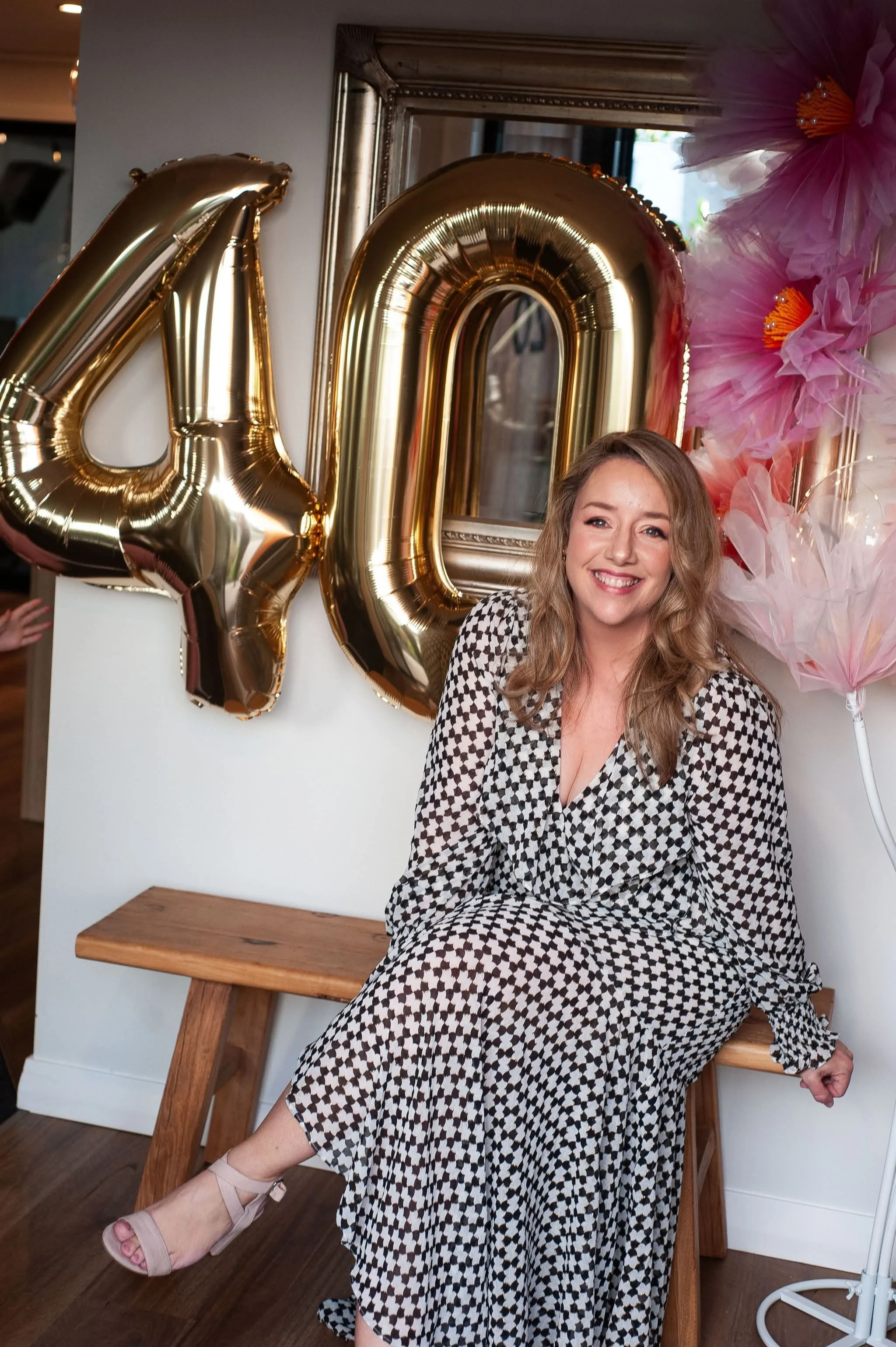 A woman with long blonde hair smiling, seated on a wooden bench, wearing a black and white patterned dress and beige high-heeled shoes, at a celebration with large gold balloons spelling '40', pink flower decorations, and a gold framed mirror in the background.