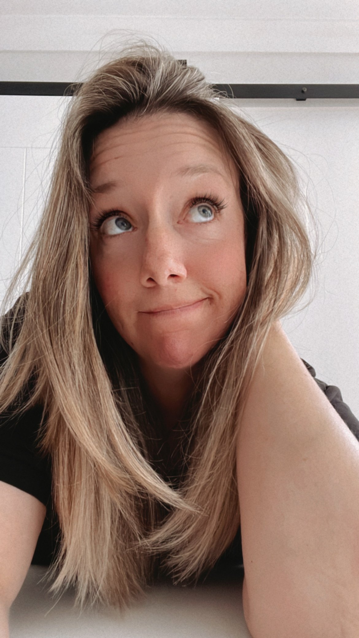 Close-up of a woman with long, light brown hair, blue eyes, and fair skin, resting her head on her arm and looking upward with a playful expression.