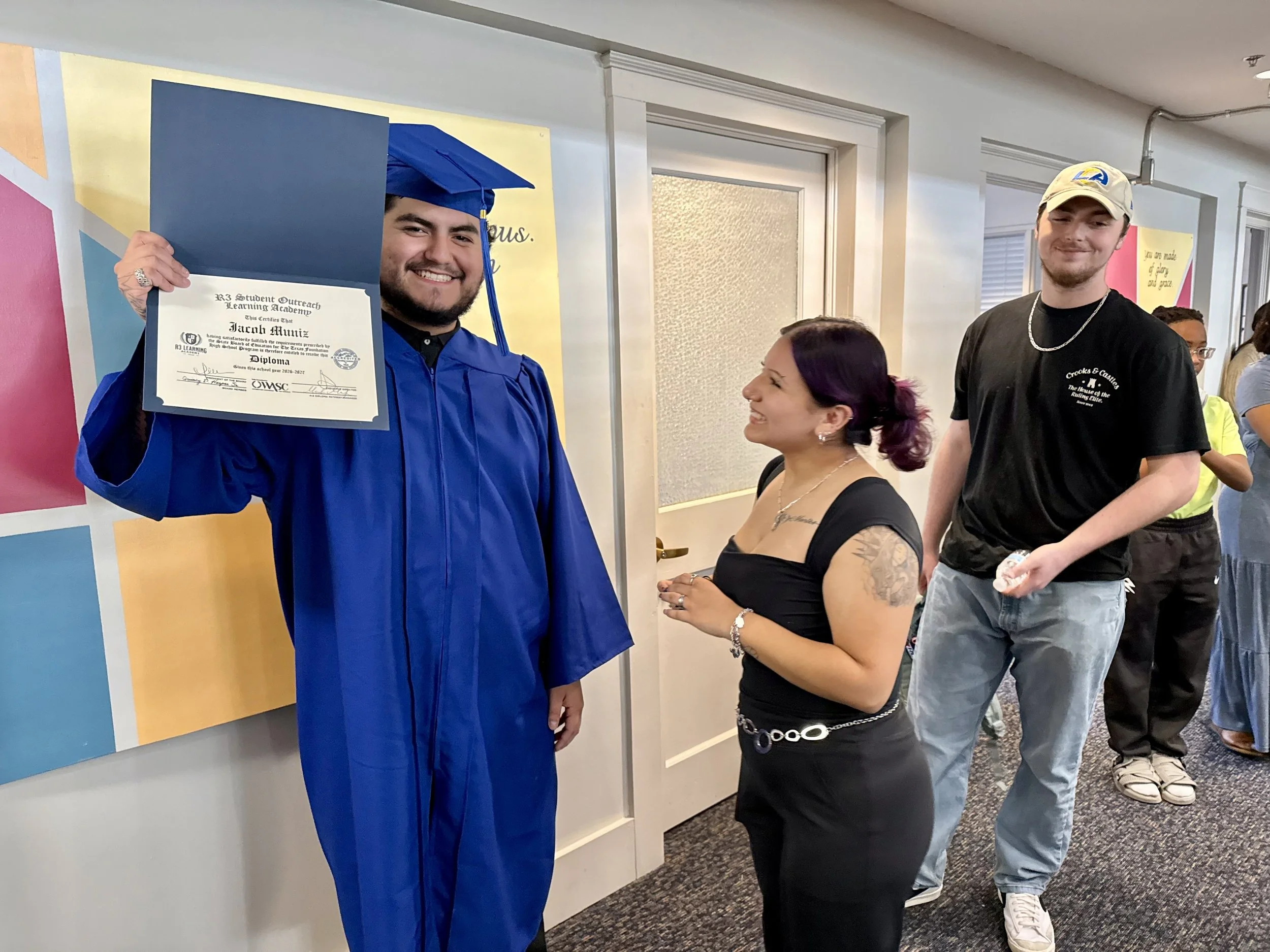 A young man in a blue graduation gown and cap is smiling and holding a diploma, standing next to a woman in black. They are smiling and hugging at a graduation ceremony in an indoor setting with wooden panels and stage lighting.