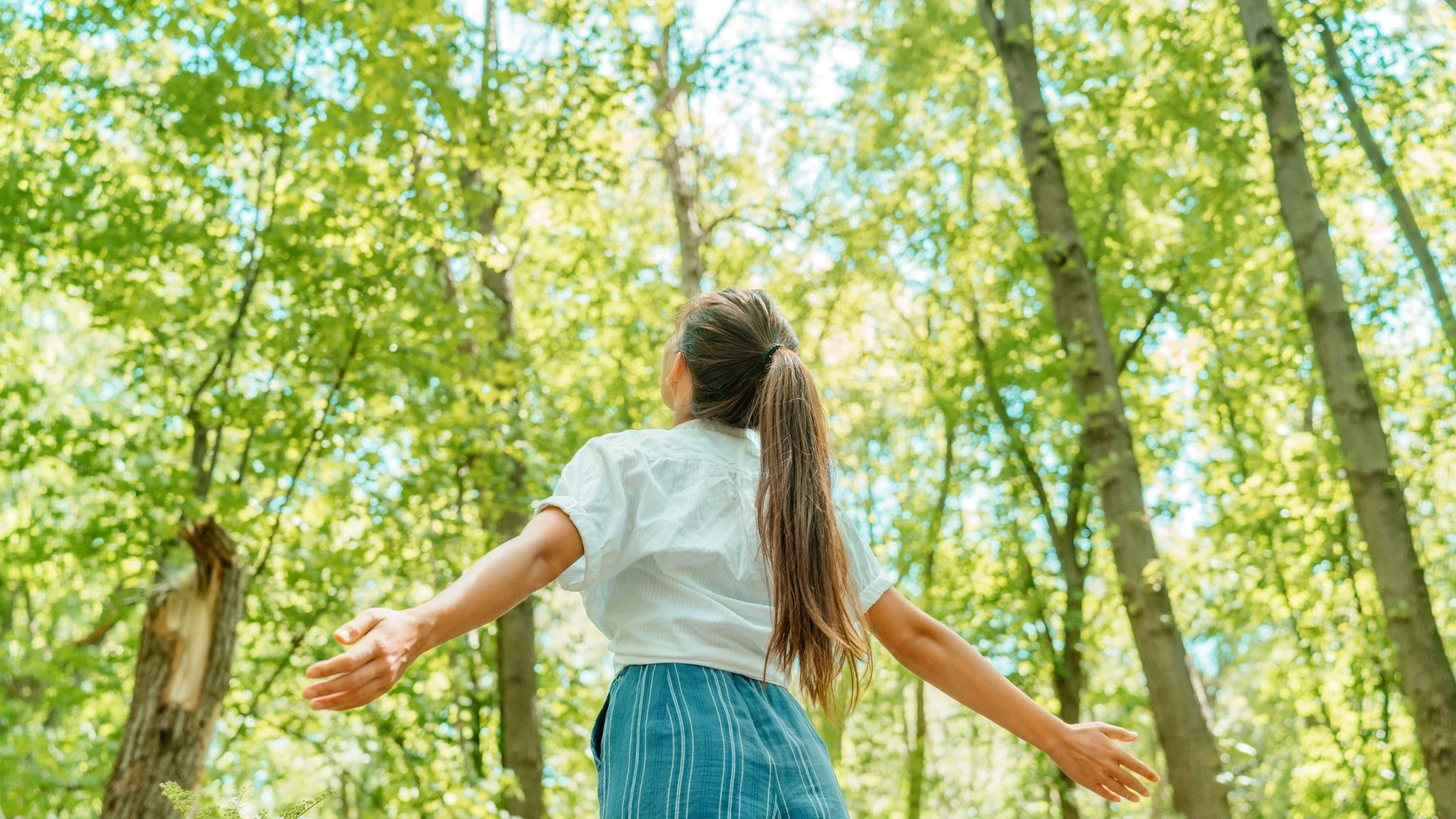 A woman with long hair tied in a ponytail, wearing a white blouse and blue skirt, standing in a lush green forest with arms outstretched.