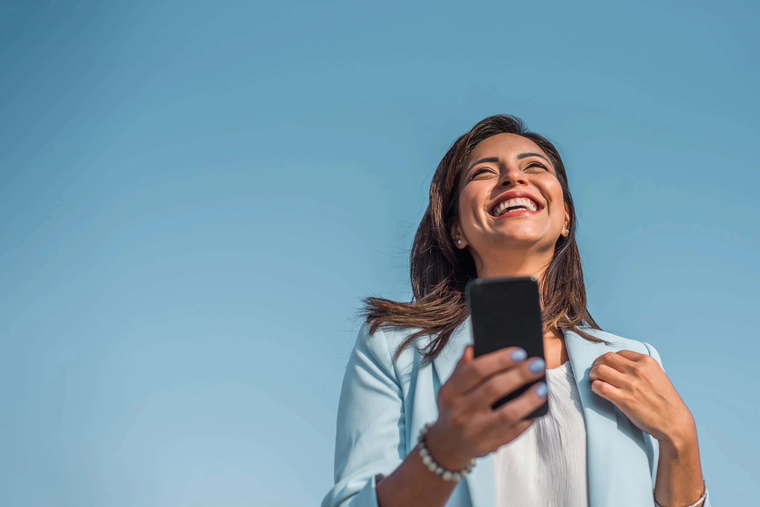 A woman smiling and holding a smartphone outdoors against a clear blue sky.