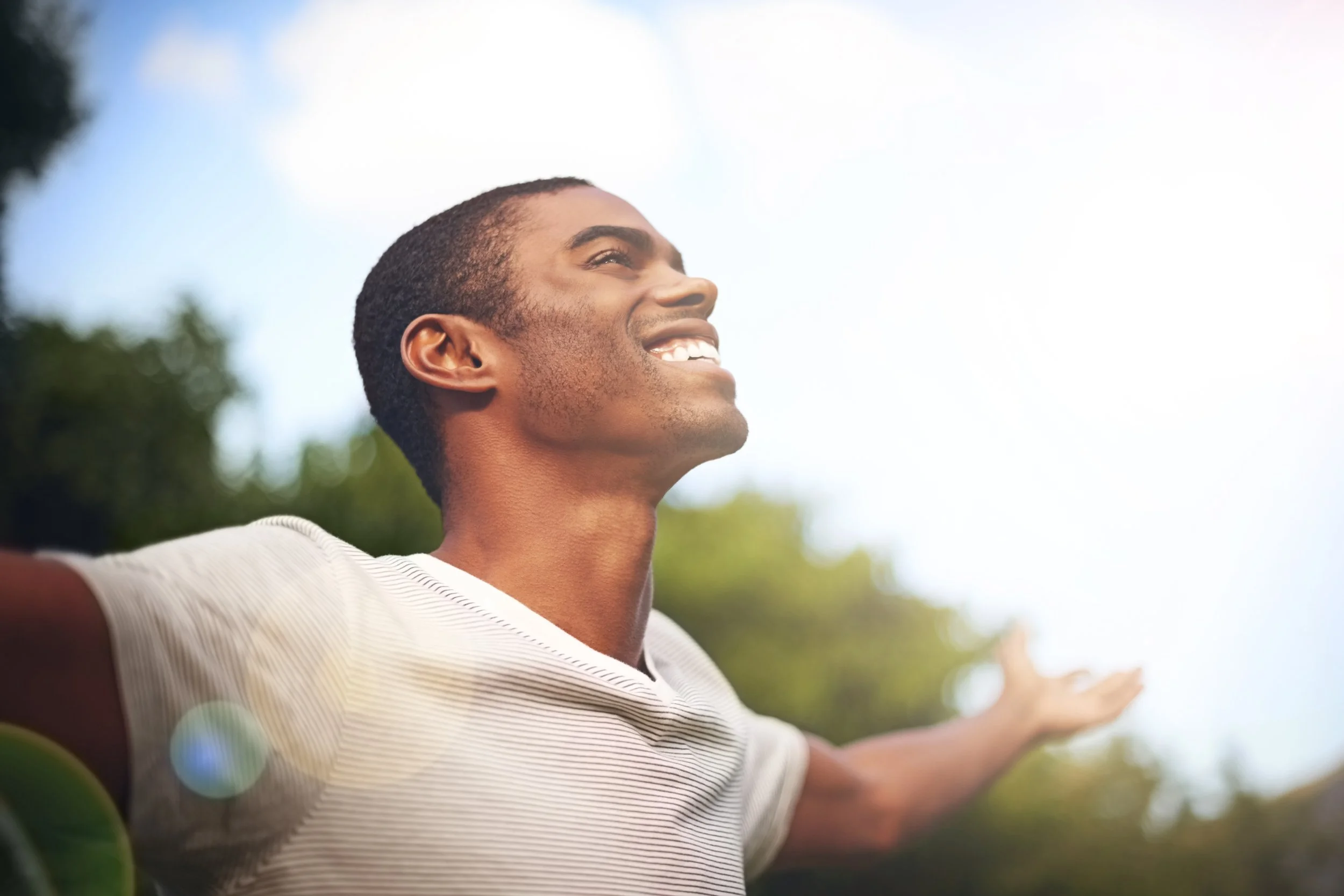 A man with arms outstretched and head tilted back, smiling, outdoors on a sunny day with trees and sky in the background.