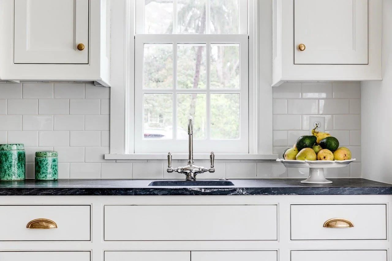 White kitchen with black marble countertop, brass drawer handles, potted plants, and a container of fruits including avocados, apples, and bananas, with a window behind the sink.