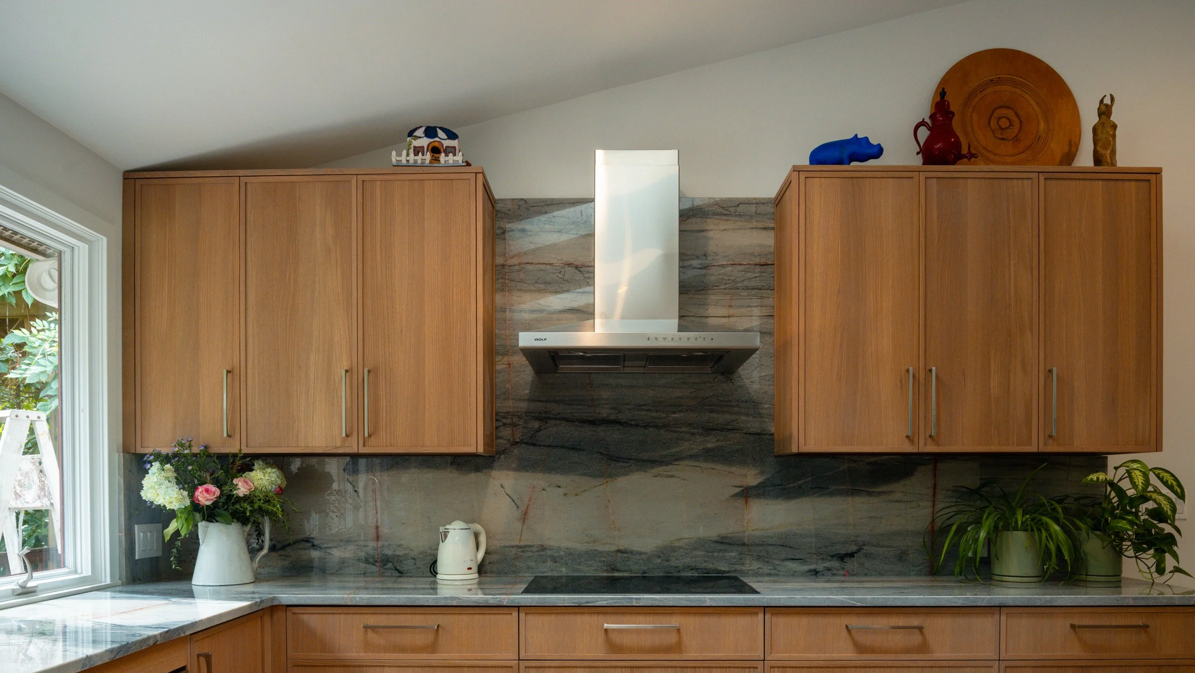 Wooden kitchen cabinets with a marble backsplash, a stainless steel range hood, potted flowers, and decorative items on top of the cabinets