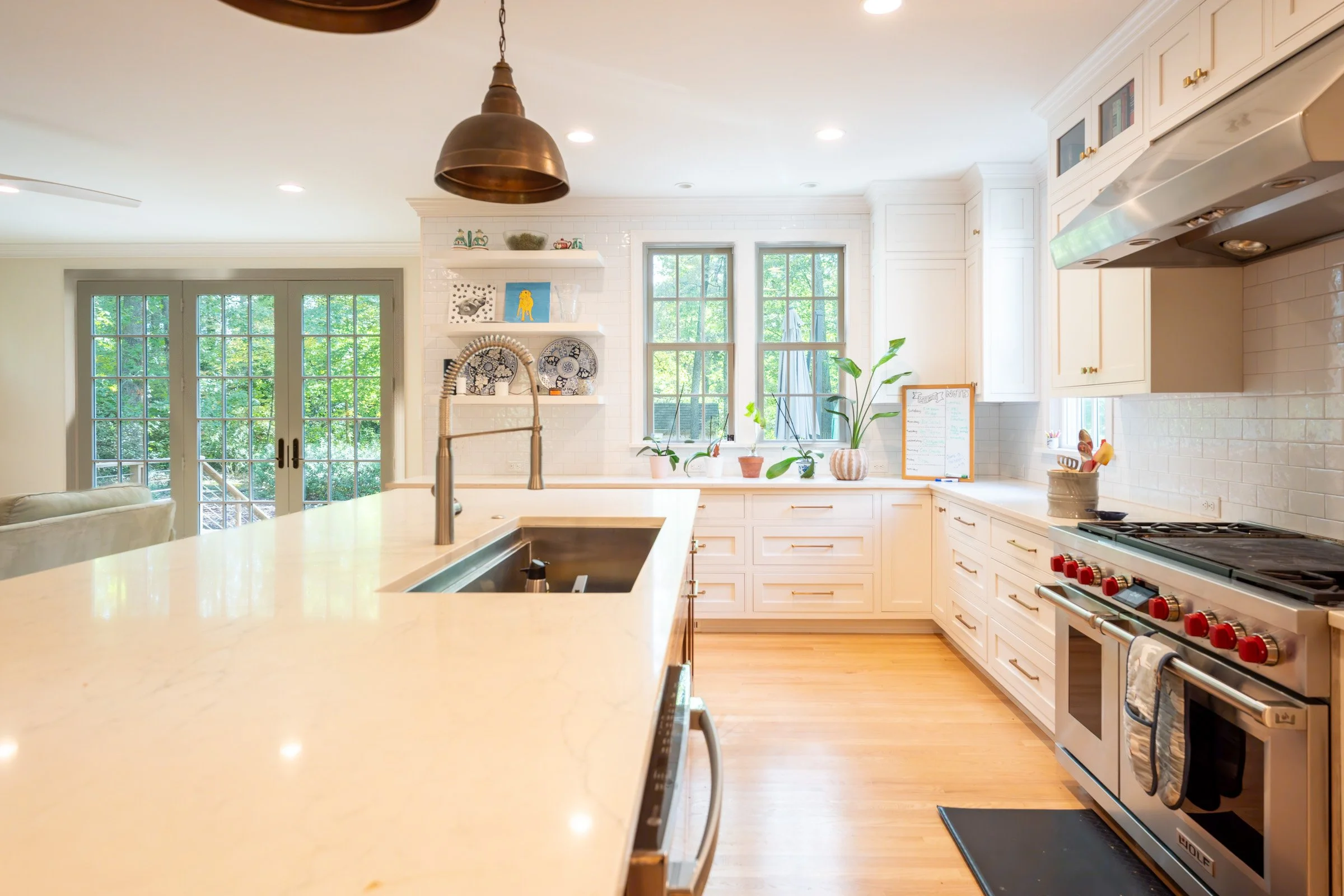 Bright kitchen with white cabinets, a large kitchen island with a built-in sink, a stainless steel stove, and multiple windows letting in natural light. Potted plants and decorative plates are on the windowsill, and a hanging pendant light is above t