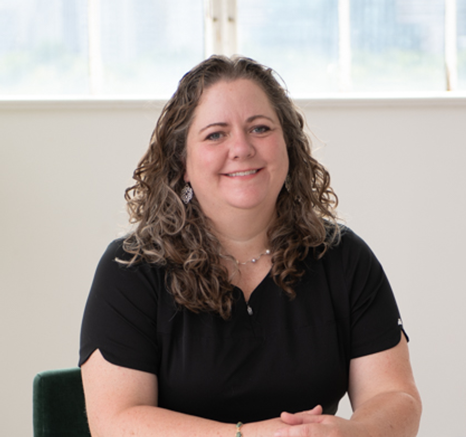 A woman with curly brown hair smiling at the camera, wearing a black shirt and jewelry, seated indoors with a window in the background.
