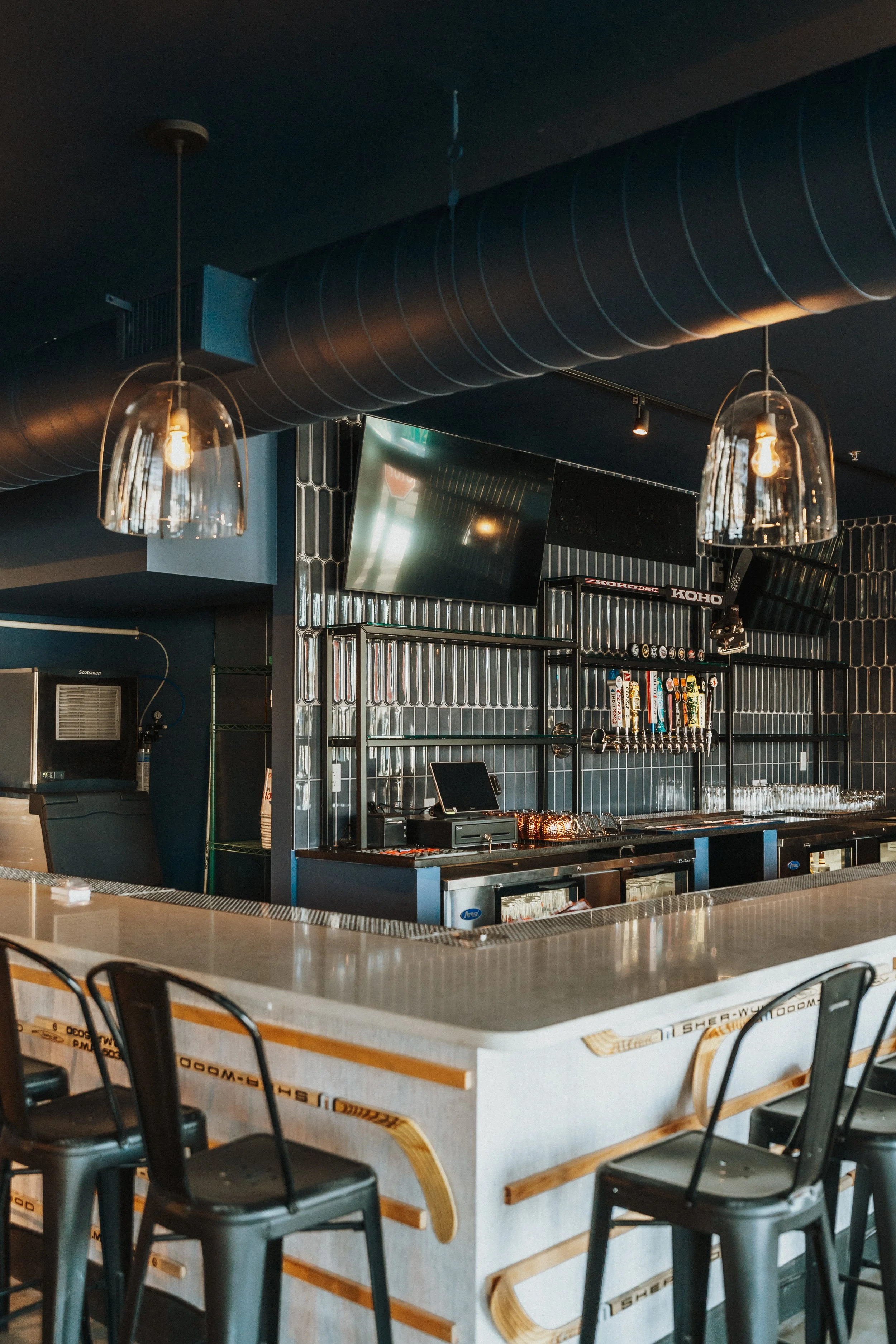 Empty bar with stools, modern industrial design, black ceiling, metallic accents, and a large flat-screen TV on the wall.