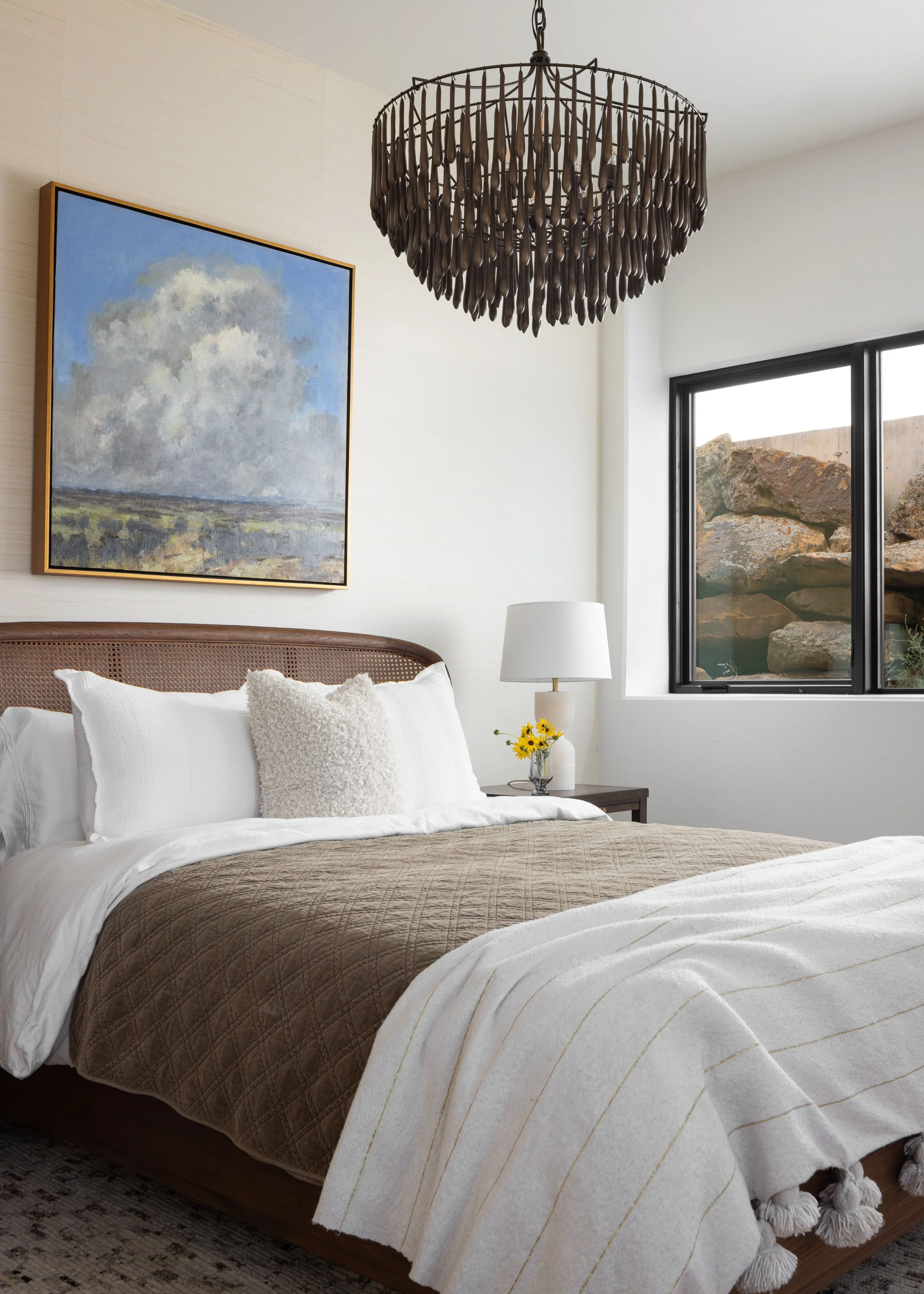 A modern bedroom with a cozy bed featuring white linens, a beige quilt, and decorative pillows. A large wall painting of a cloudy sky hangs above the bed. A stylish chandelier is mounted on the ceiling, and a window with a view of rocks allows natural light to enter. A bedside table with a lamp and a small vase of yellow flowers completes the room's decor.