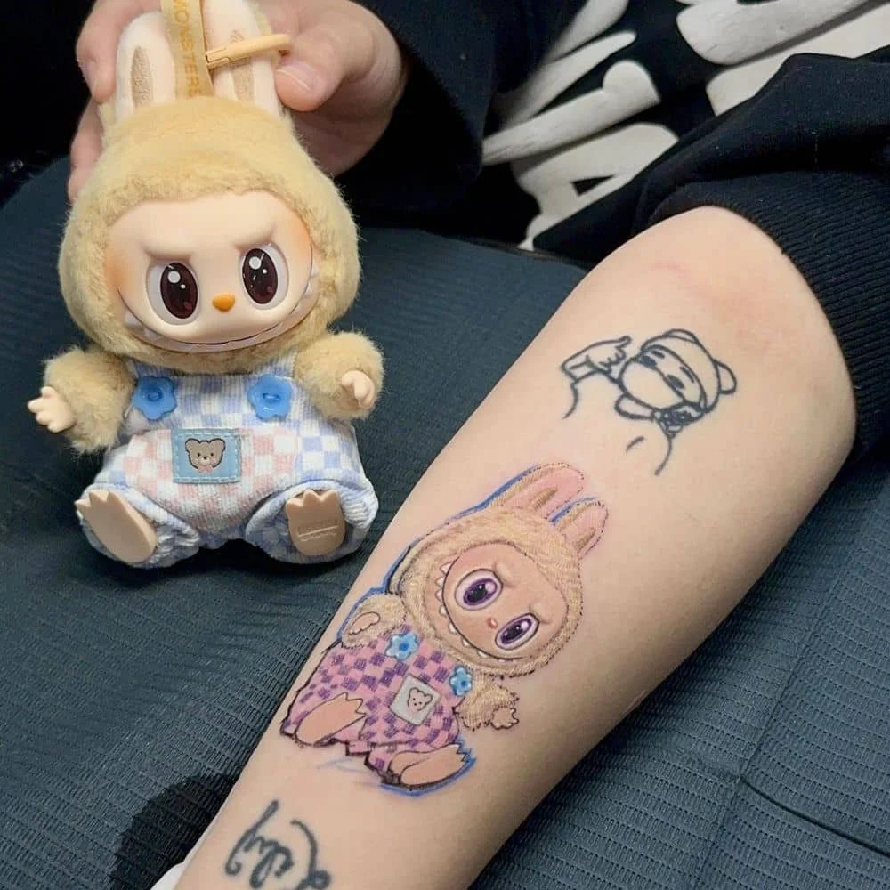 A person's arm with a colorful tattoo of a character with big eyes and rabbit ears, wearing a checkered outfit with floral details. Next to the tattoo, there is a small line drawing of the same character. The person is holding a plush toy of the same