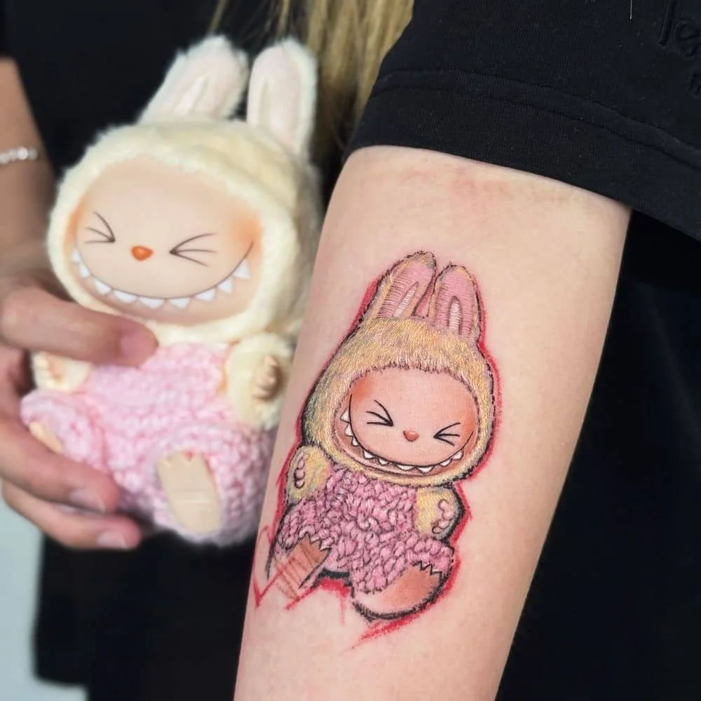 A person's arm with a colorful tattoo of a character with big eyes and rabbit ears, wearing a checkered outfit with floral details. Next to the tattoo, there is a small line drawing of the same character. The person is holding a plush toy of the same