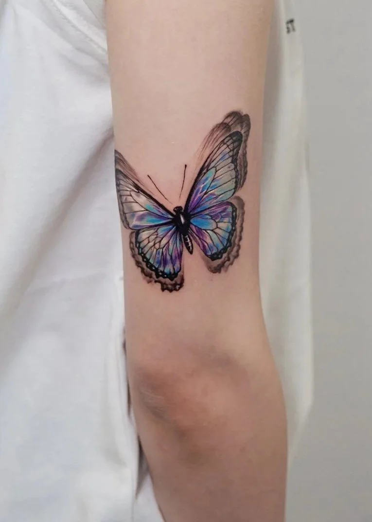 Close-up of a person's arm with a colorful butterfly tattoo on the upper arm, wearing a white sleeveless shirt.
