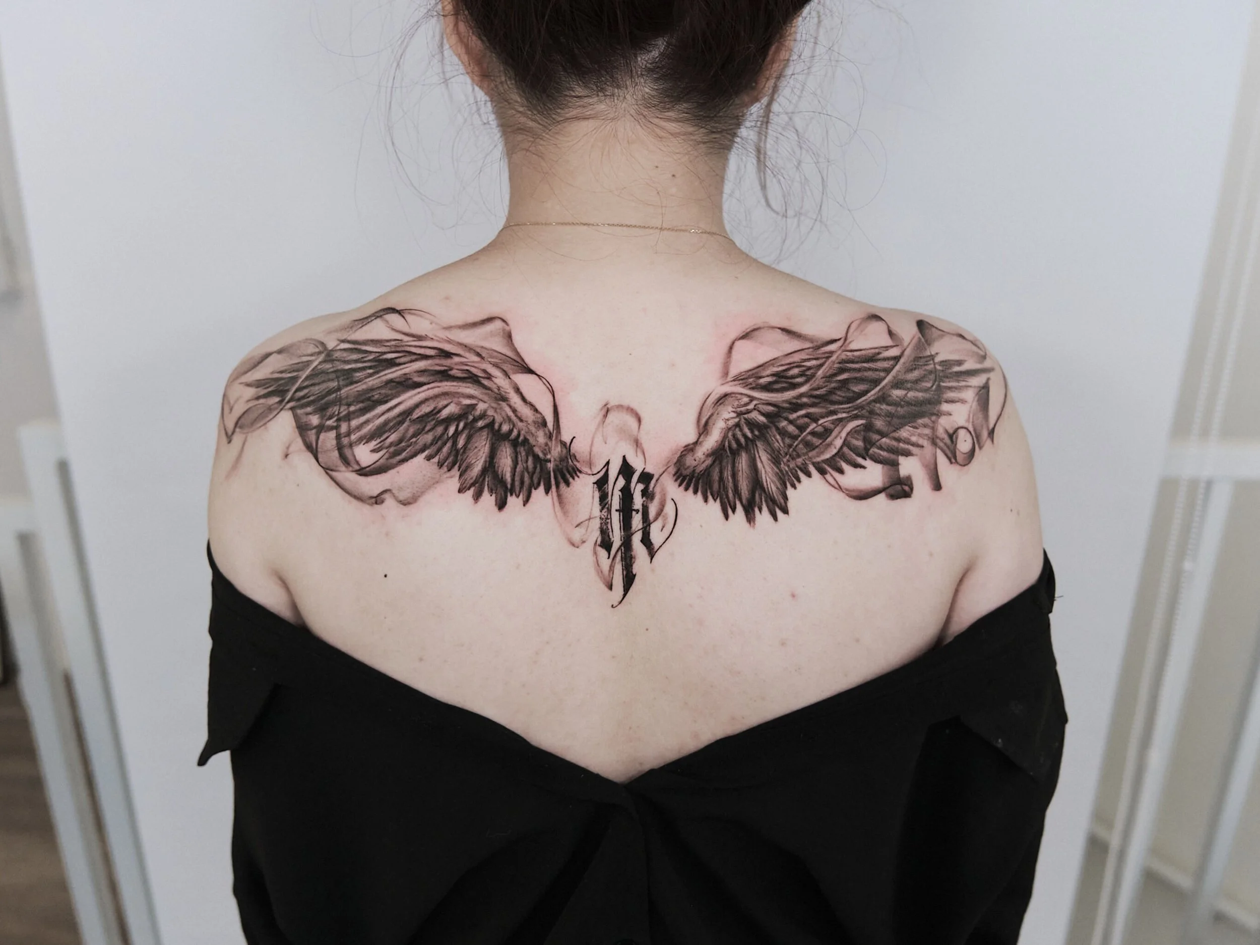 Tattoo of black shaded angel wings with a stylized letter 'H' in the center on a person's upper back and shoulders.