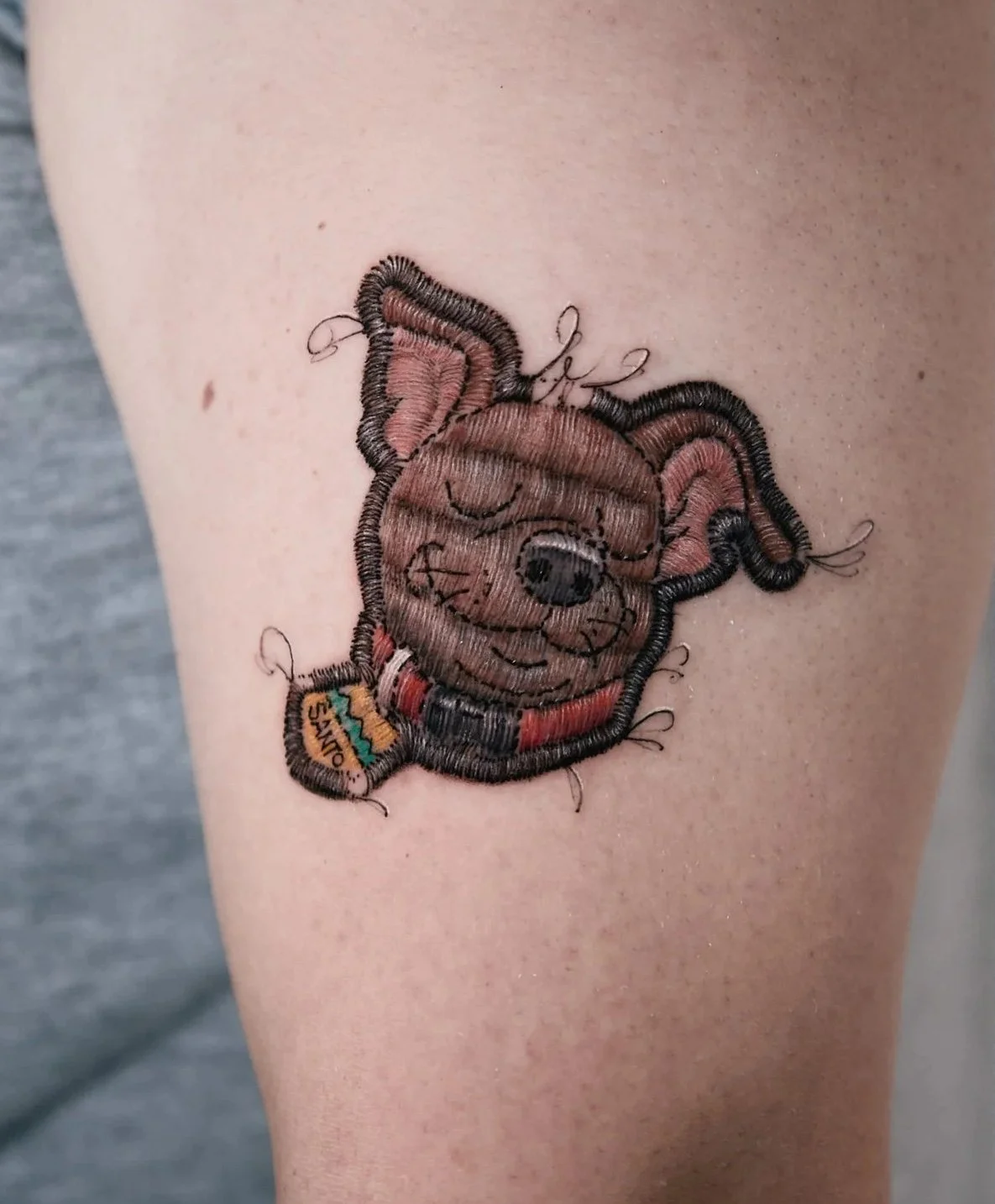 Close-up of a person's upper arm with a detailed embroidered teddy bear tattoo wearing a rainbow-colored accessory and a small ribbon labeled 'Stinky'.