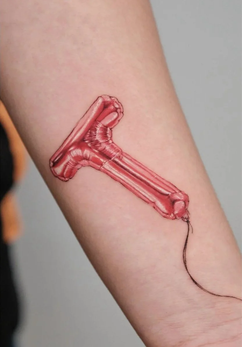 Close-up of a person's arm with tattoos, including a fingerprint with the year 1989 and a heart shape, and a red microphone tattoo, with the person wearing a black shirt and jeans.