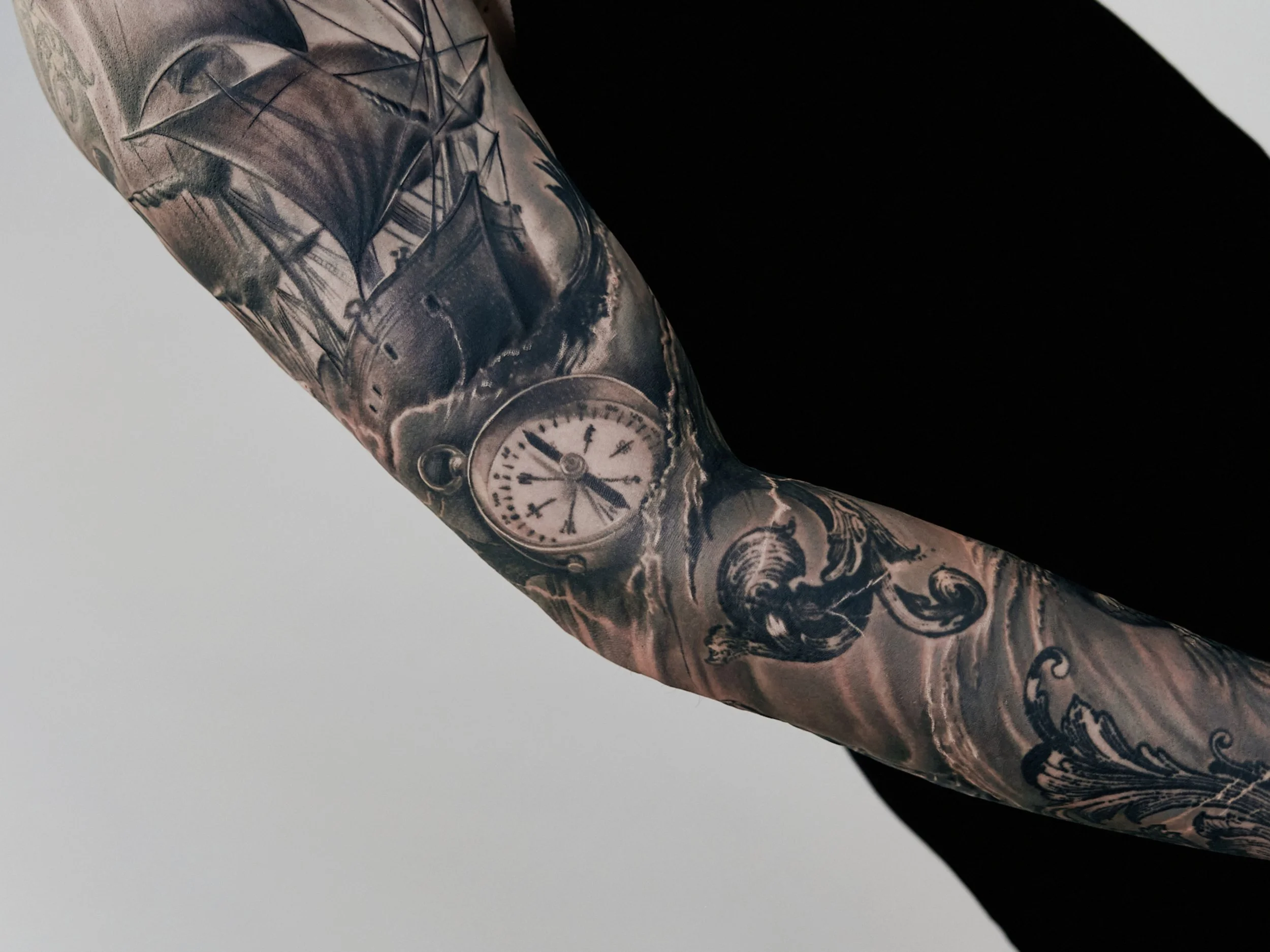 Close-up of a person's arm with a detailed black and gray tattoo of a sailing ship, a pocket watch, and swirling waves.