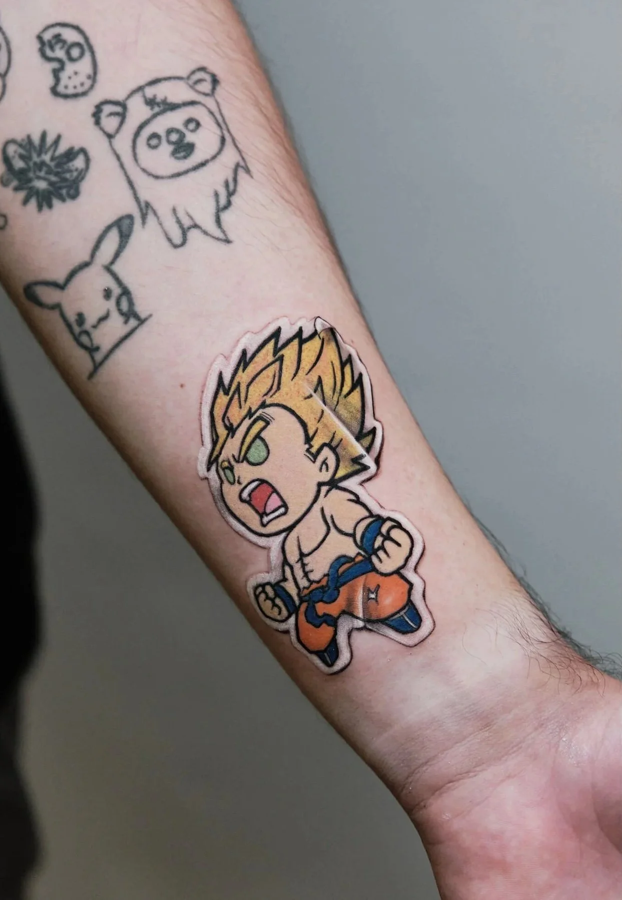 A person's arm with multiple tattoos, including a cartoon character with spiky hair and an angry expression, a pig, a lion's face, and other small designs. The person is wearing a black shirt.