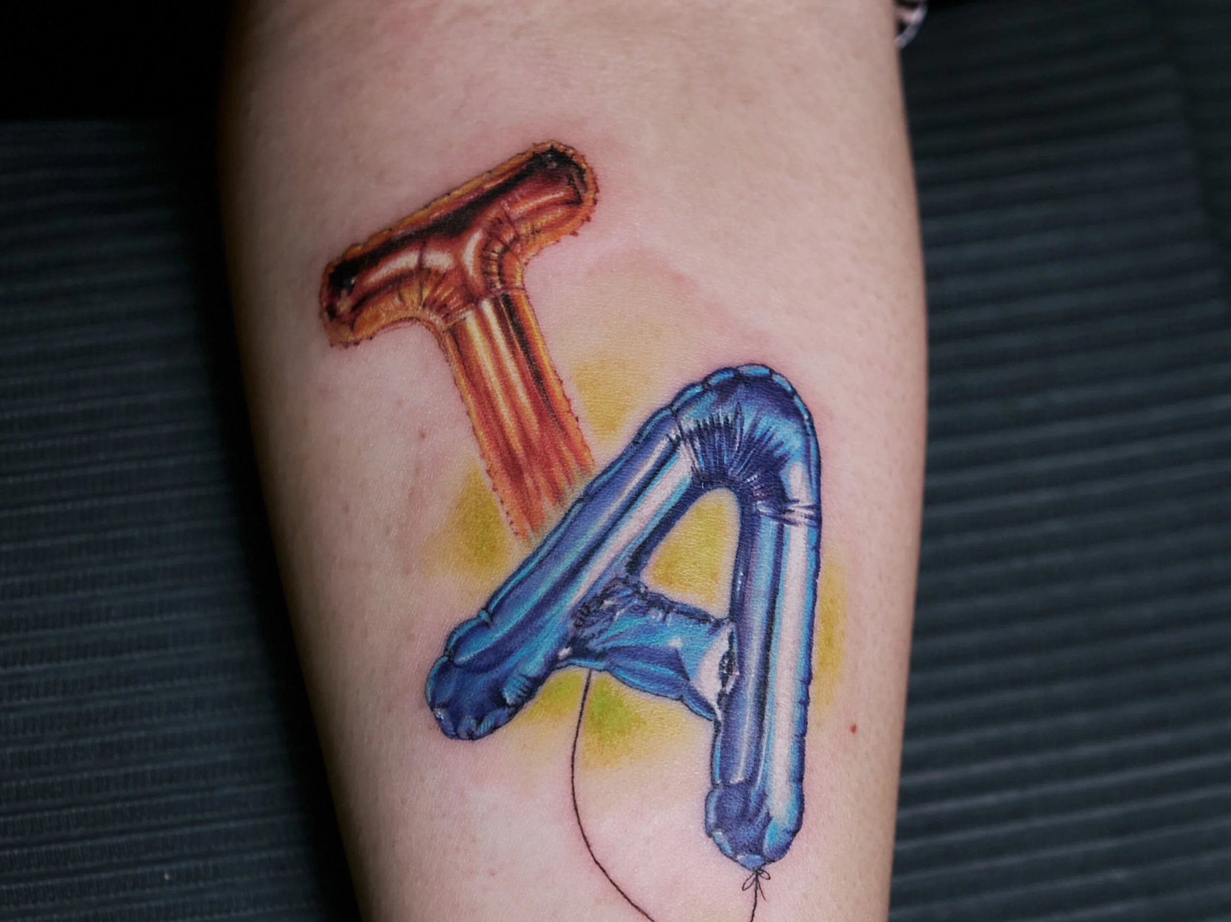 Colorful tattoo of a balloon shaped like the letter A on person's arm.