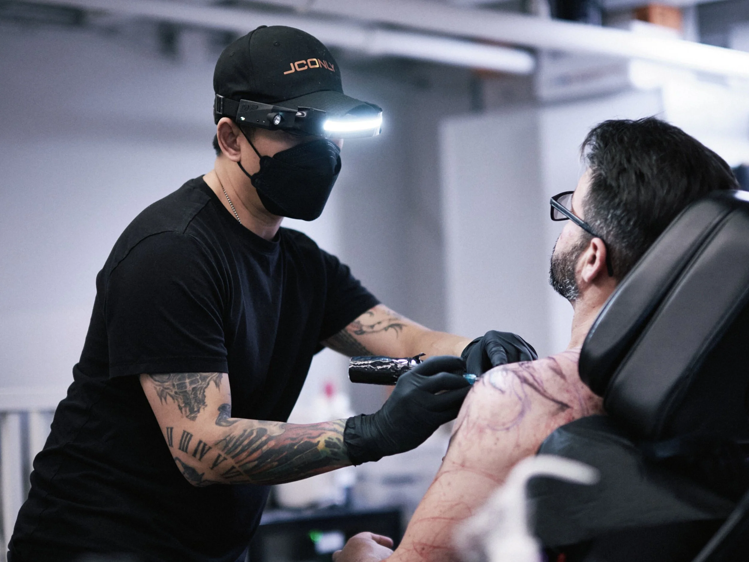 A tattooed man is sitting in a chair receiving a tattoo on his shoulder while a tattoo artist, wearing a black mask, gloves, and a headlamp, works on his skin in a tattoo studio.