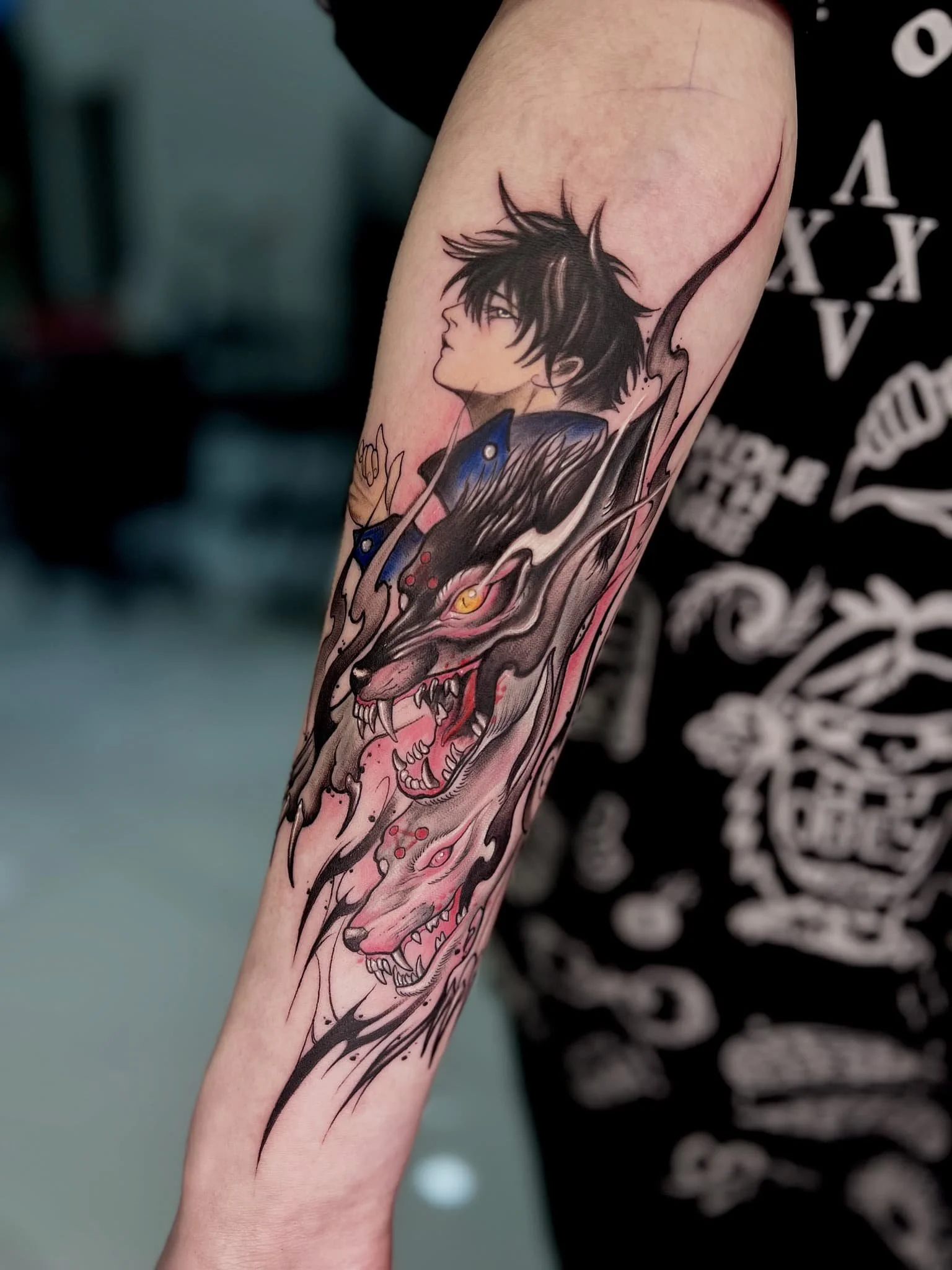 Black and grey tattoo on forearm featuring a young man with styled hair and a fierce dragon with multiple heads, sharp teeth, and detailed features.