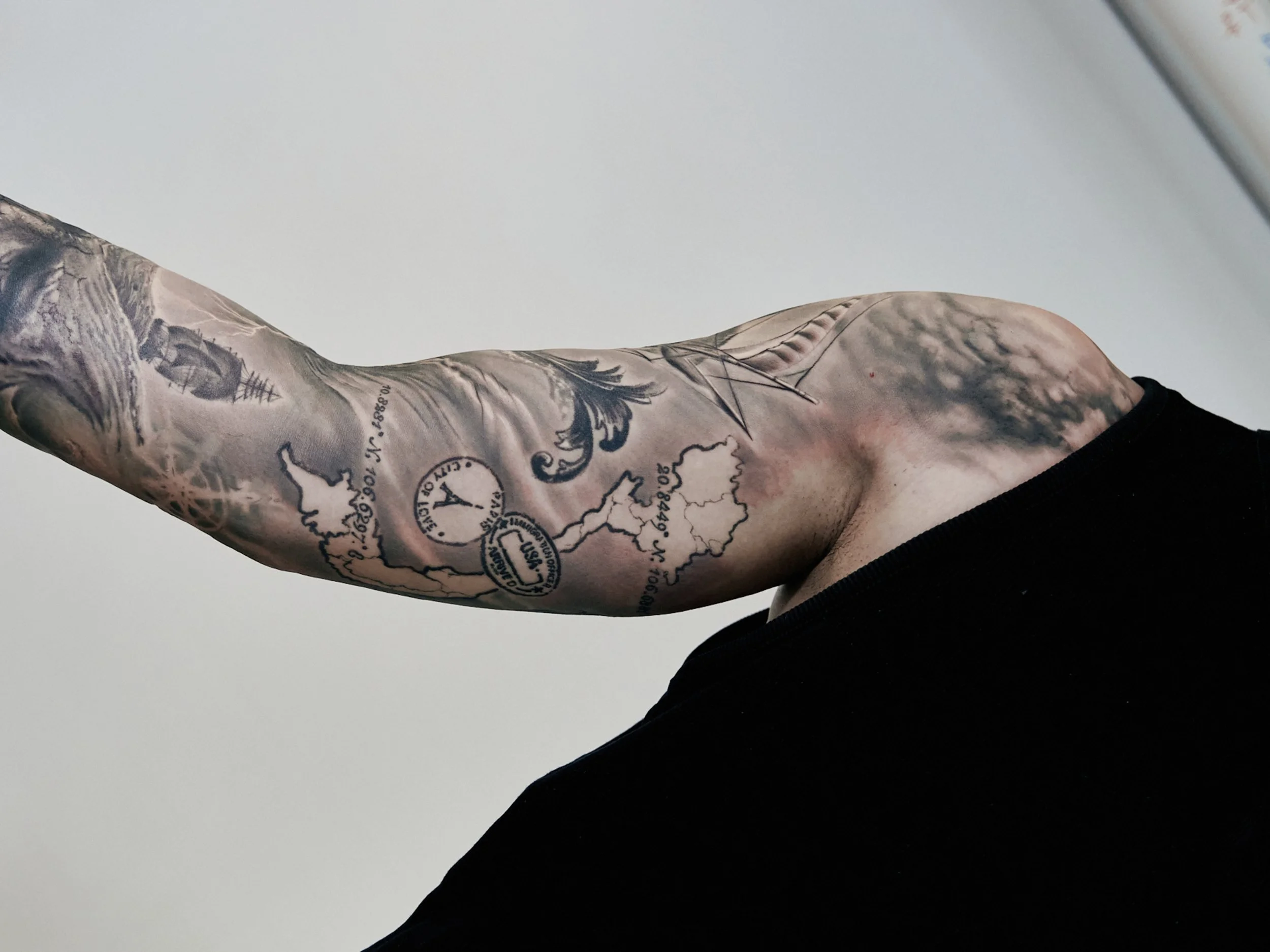 Close-up of a person's upper arm with detailed black and gray tattoos including a clock, map outline, and various designs, against a plain white background.