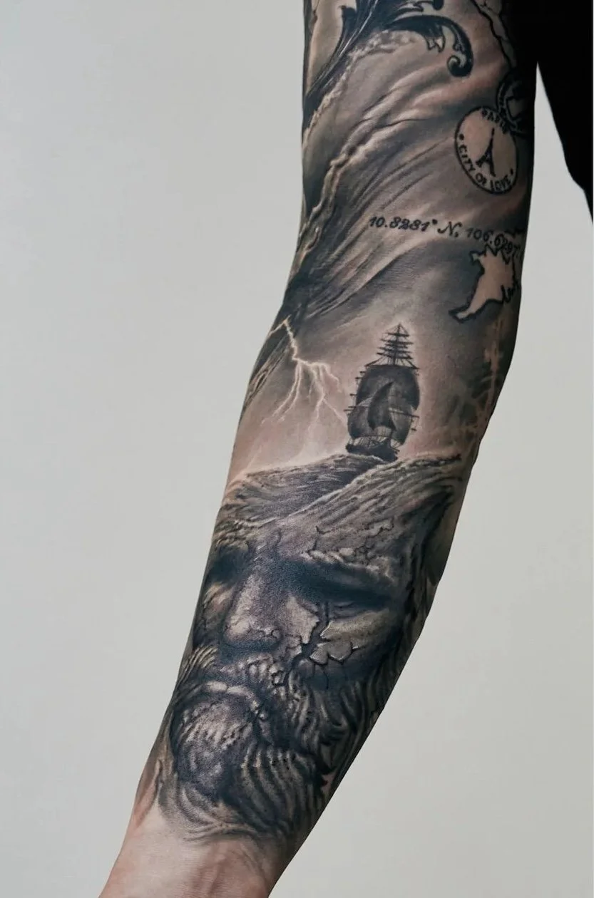 Close-up of a detailed black-and-grey tattoo sleeve featuring a bearded stone god, a tall sailing ship on stormy seas with lightning, geographic coordinates, and a Paris passport stamp.