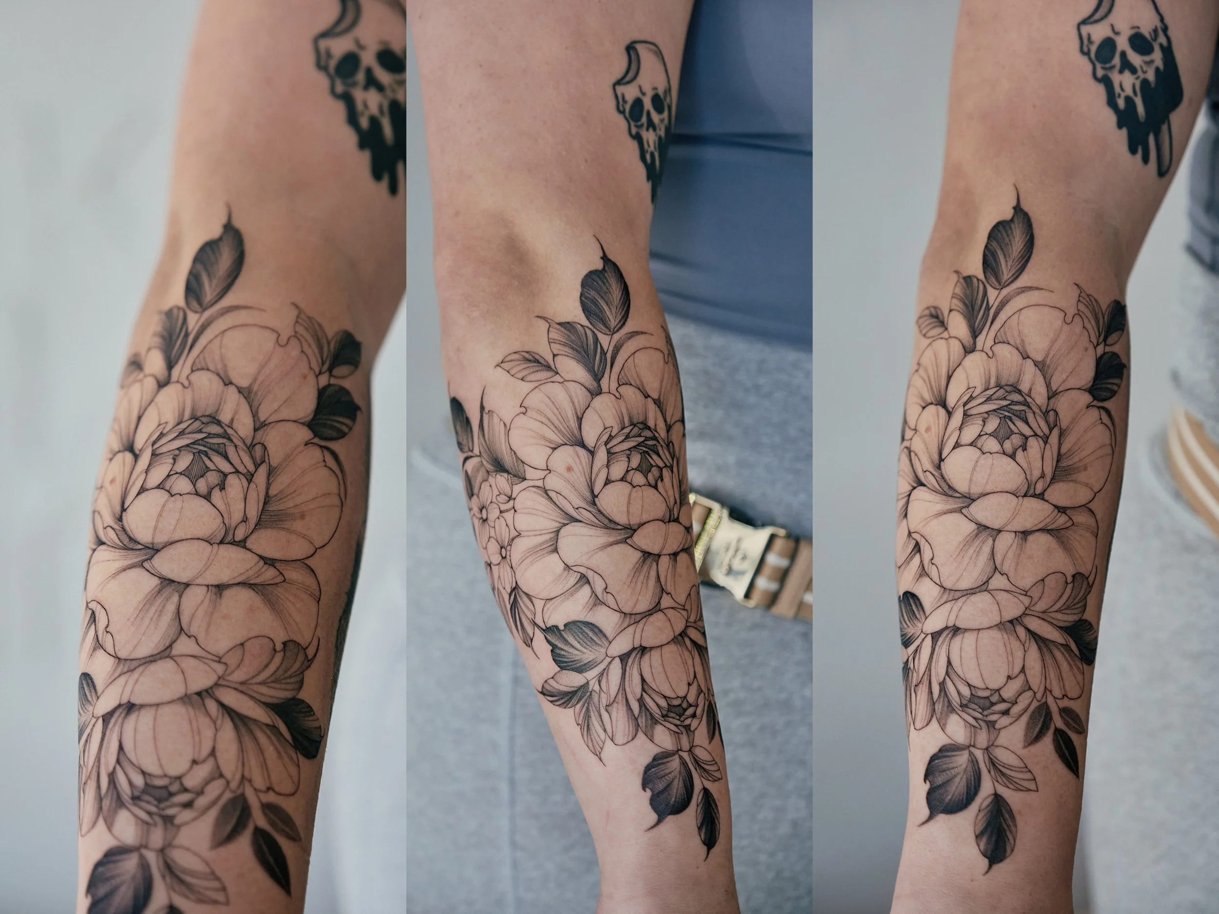 A person's forearm with a large black and grey floral tattoo featuring detailed peonies and leaves.