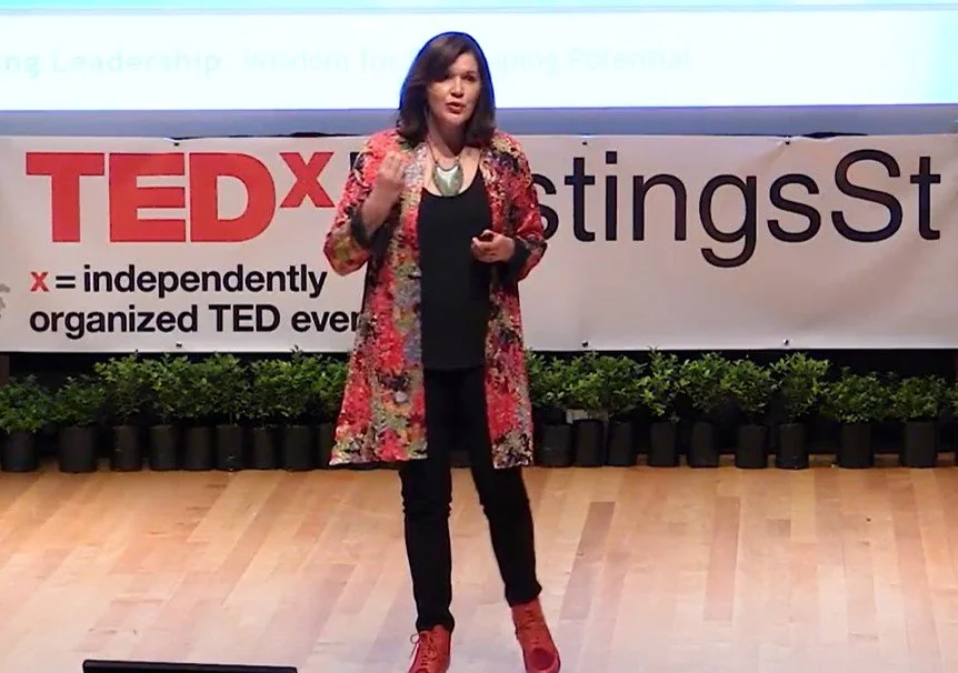A woman speaking on stage at a TEDx event, wearing a colorful blazer, black top, black pants, and red boots, with a sign behind her that reads 'TEDx' and 'StingsSt' and potted plants arranged along the back of the stage.