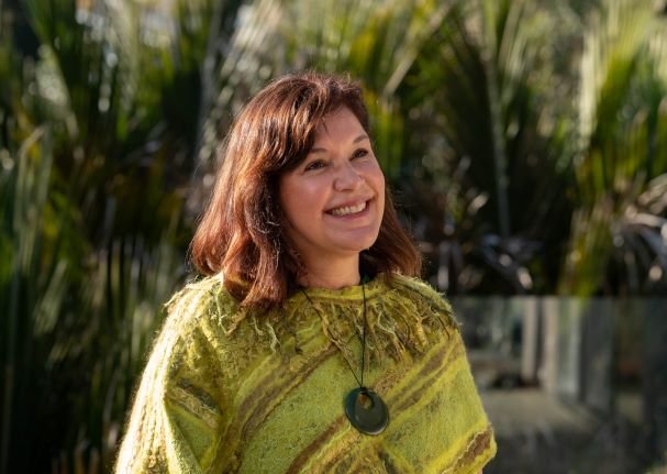 A smiling woman with brown hair wearing a yellow-green patterned poncho and a black necklace, outdoors with green foliage in the background.