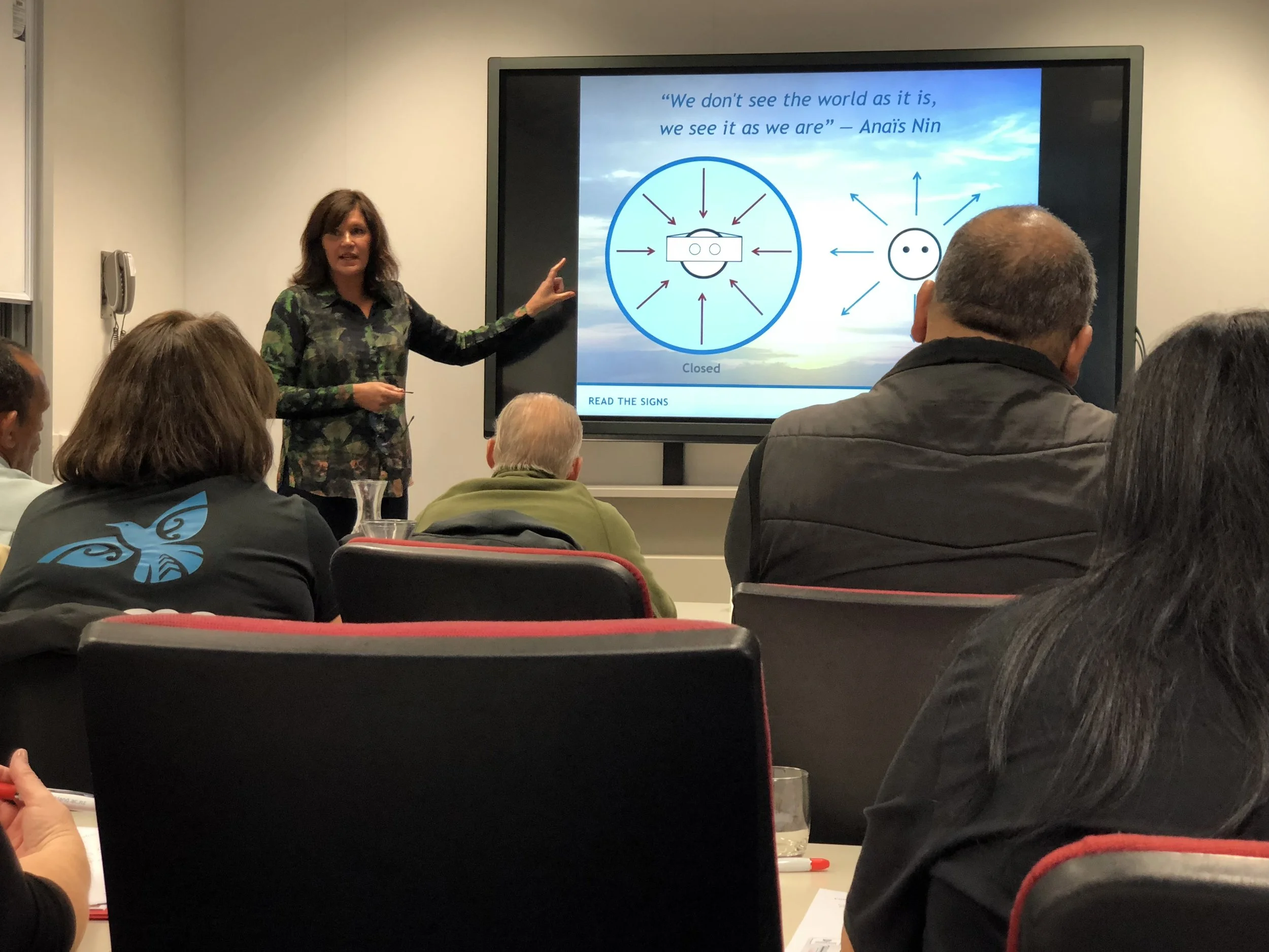 A woman in a dark, floral shirt is pointing at a large screen displaying a quote and diagrams during a presentation. The slide shows a quote by Anaïs Nin about perception, with illustrations of a smiling face wearing virtual reality goggles, and a simple face with dotted eyes and arrows pointing outward.