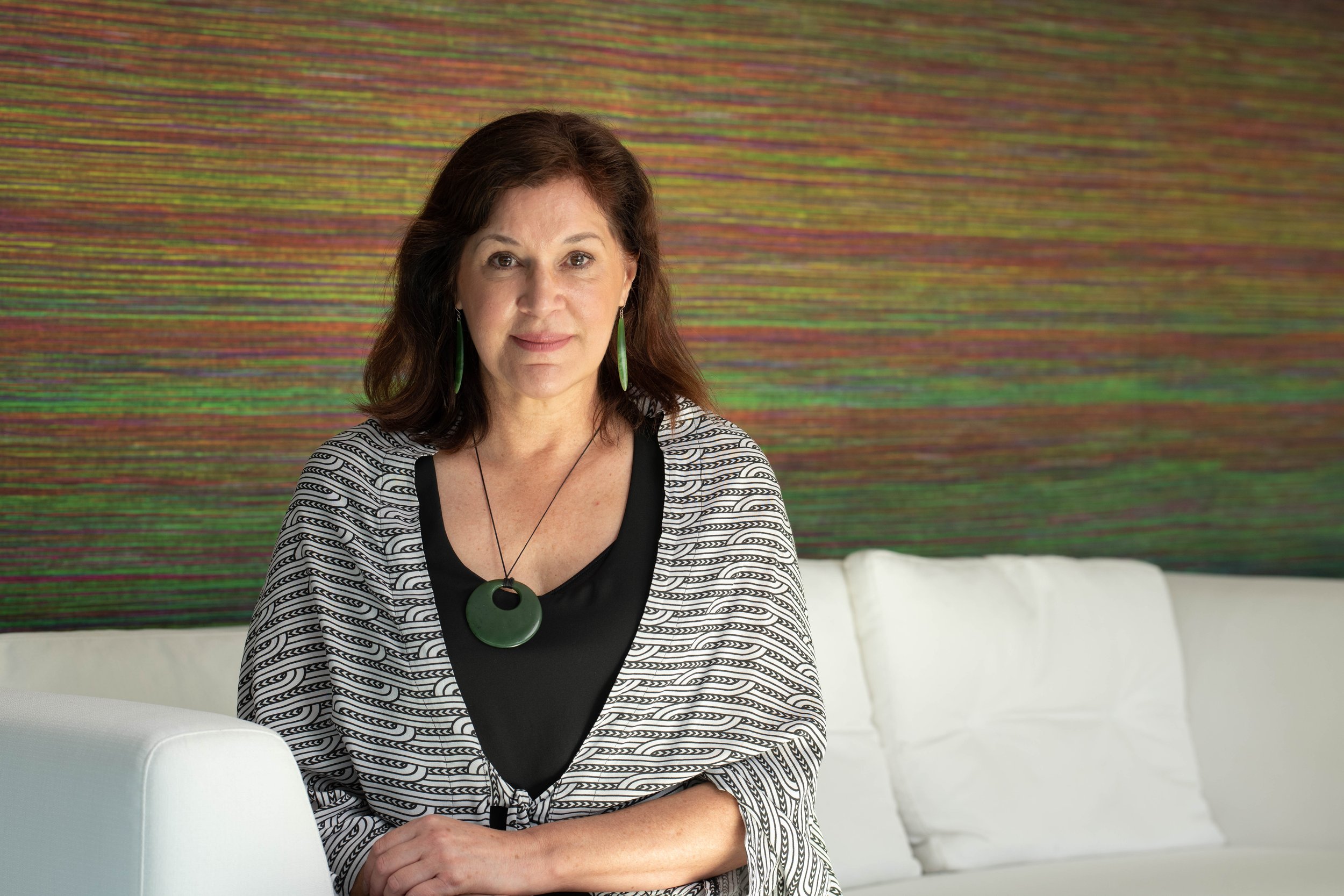 A woman with brown hair wearing green jewelry, a black top, and a chevron patterned jacket, sitting on a white sofa in front of a multicolored abstract artwork.