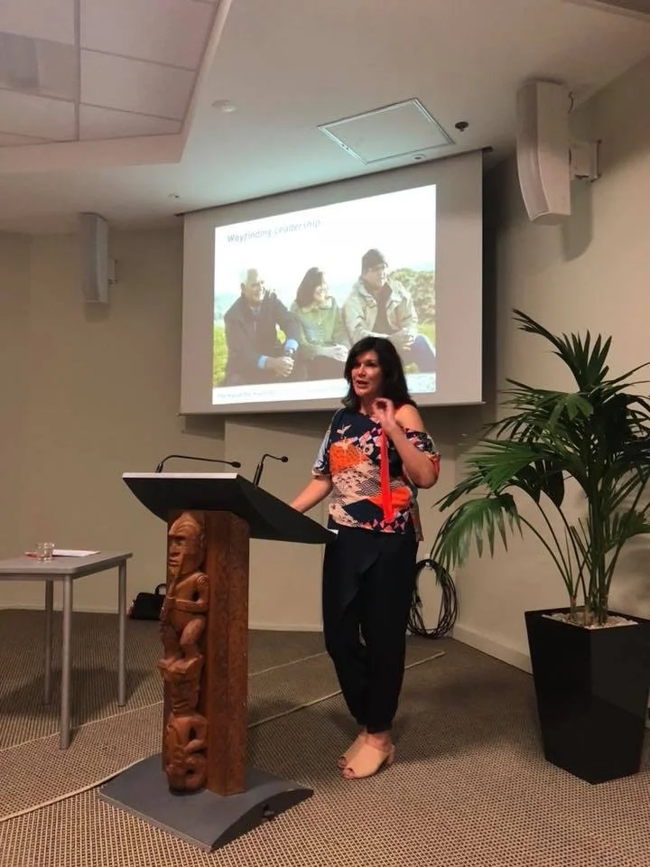 Woman speaking at a podium during a presentation in a conference room, with a slide showing a group of three people outdoors projected behind her, and a large potted plant on her right.