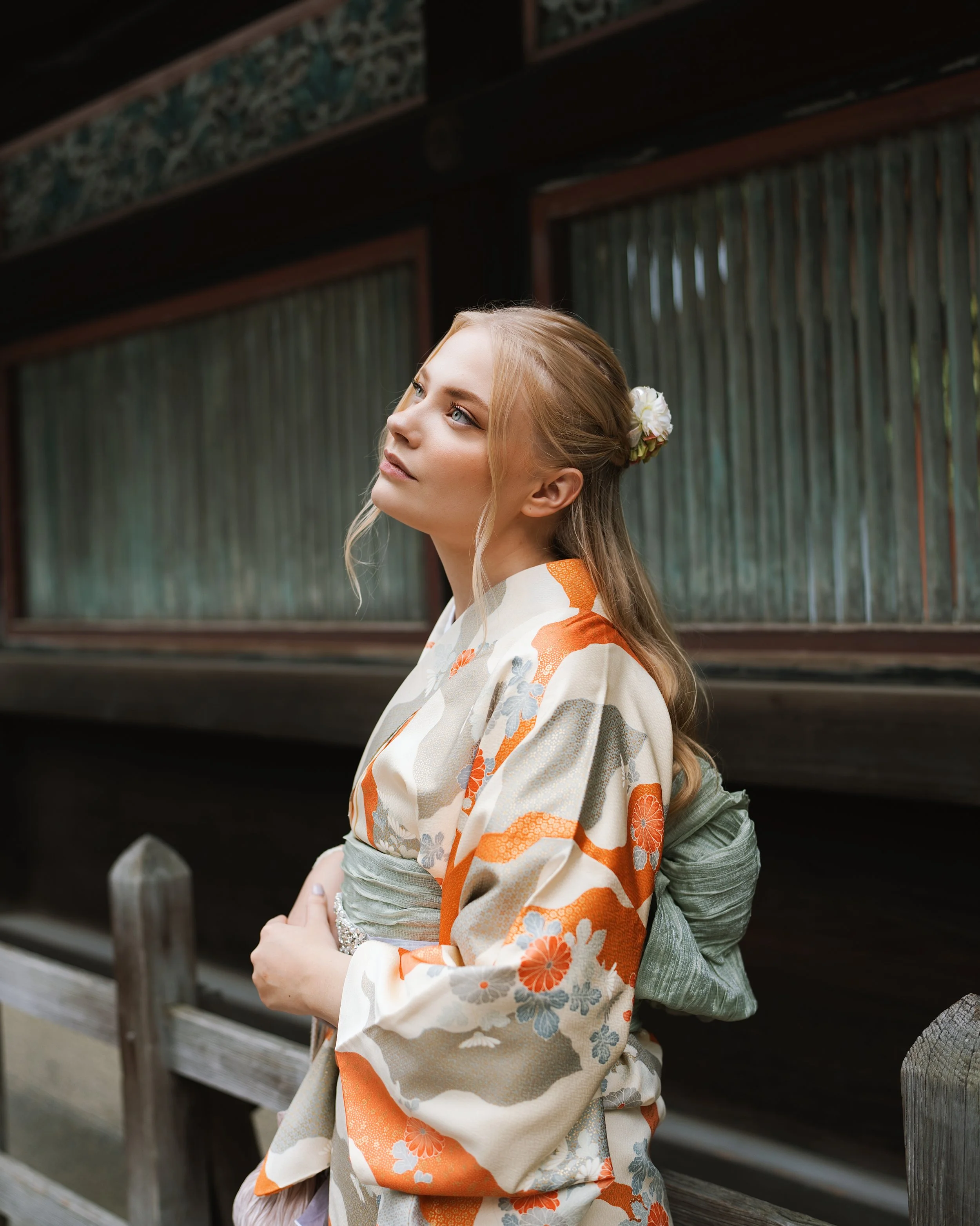 A young woman wearing a traditional Asian kimono with orange, grey, and white floral patterns, standing outdoors near wooden structures, with a flower in her hair.